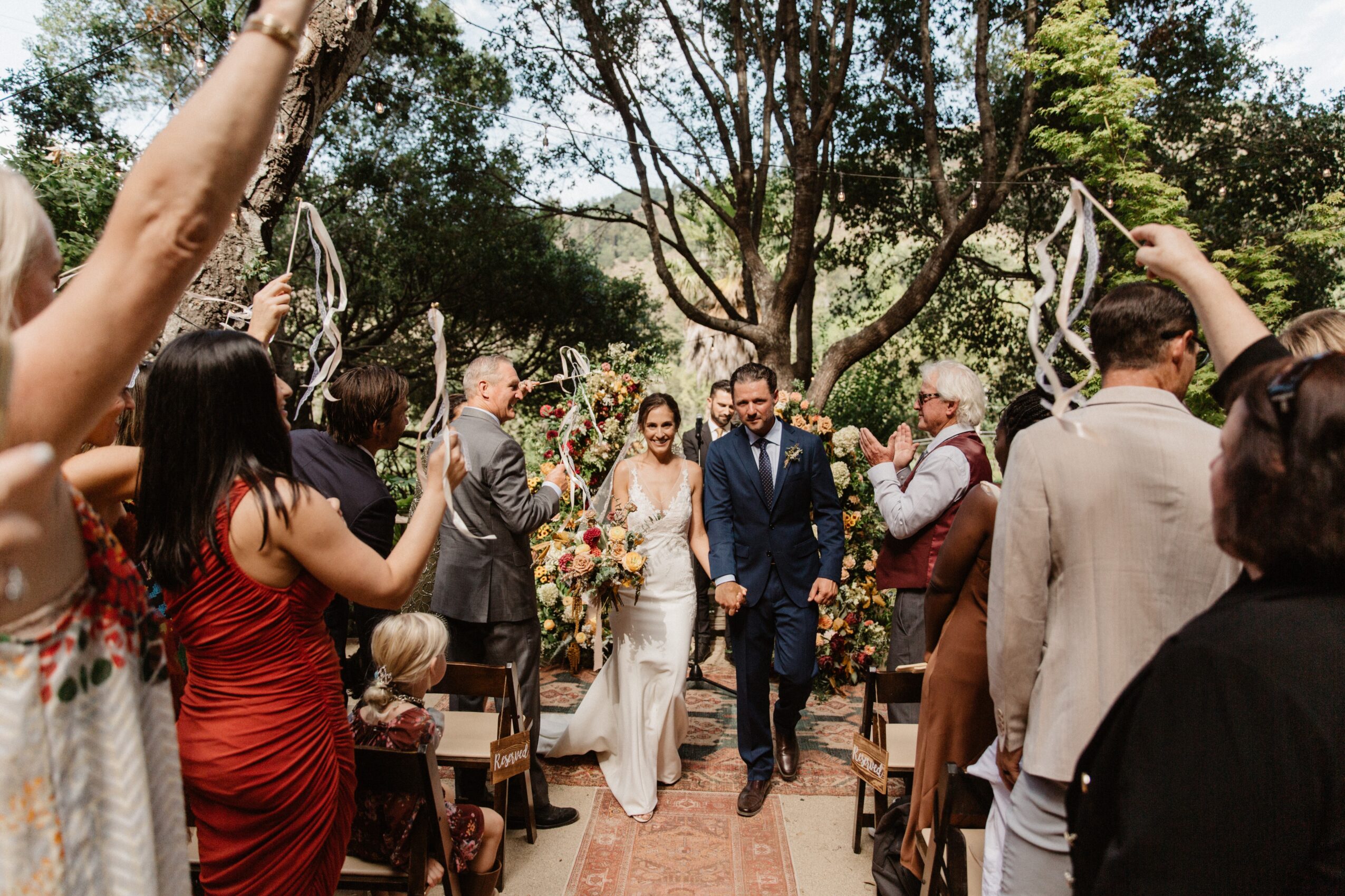 Bride and groom walking down an outdoor aisle surrounded by guests and trees, celebrating wedding.