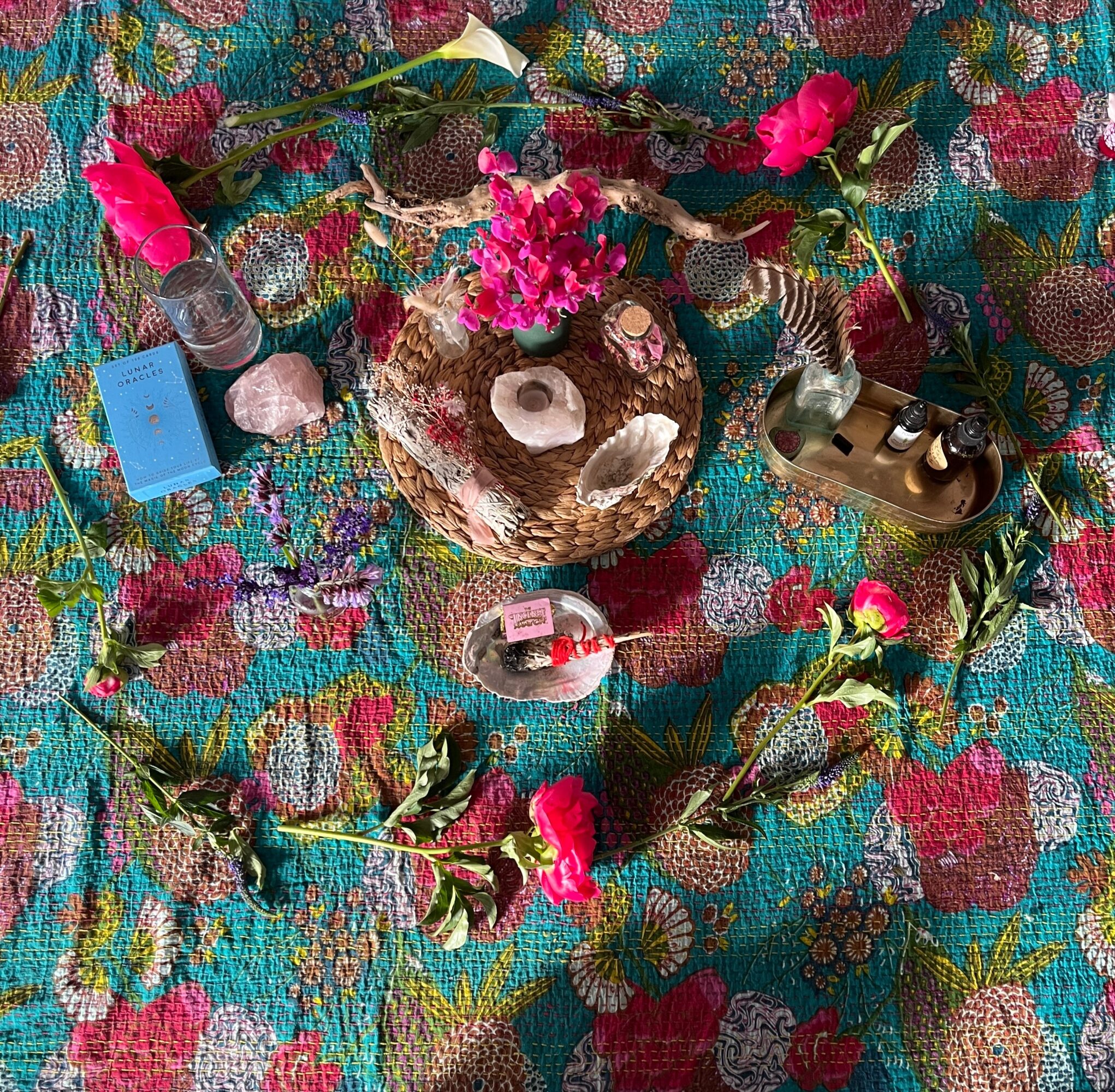 Decorative arrangement with pink flowers, a small bottle, a pink card, a shell, and a vintage camera on colorful fabric.
