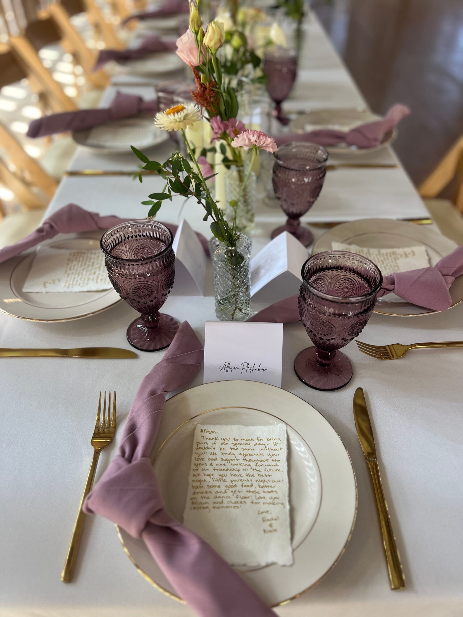 Table setting with plates, purple glasses, gold utensils, pink napkins, and a floral centerpiece.
