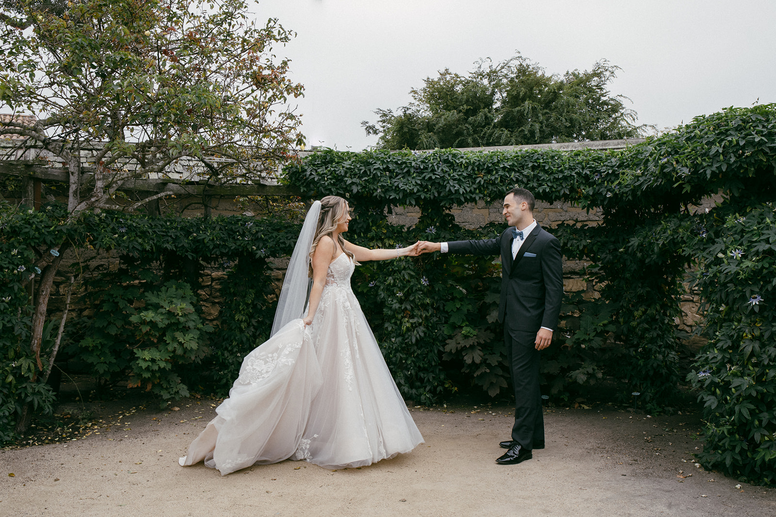 Bride and groom holding hands outdoors with greenery background, bride in wedding dress and veil, groom in tuxedo.