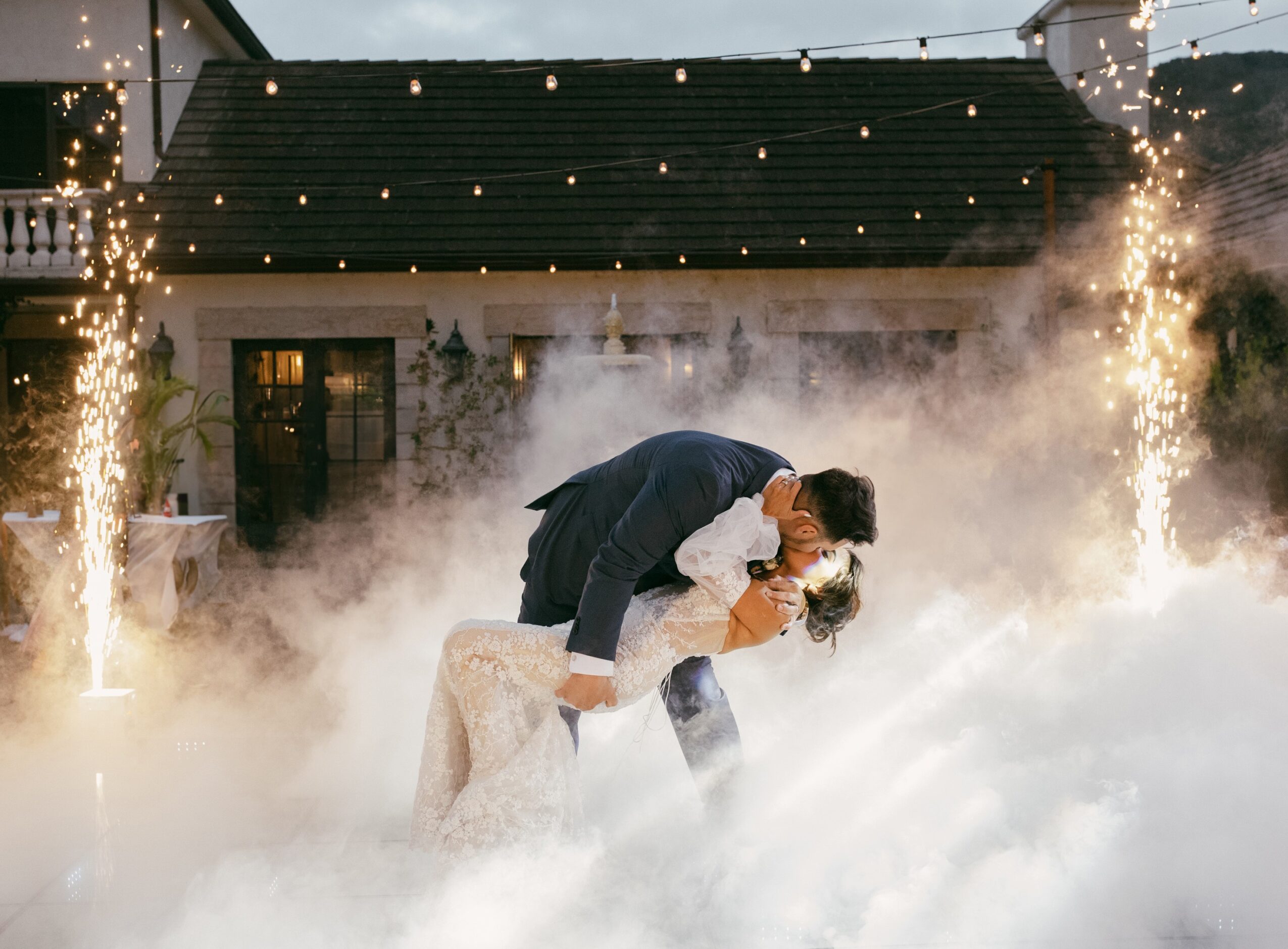 Couple dancing outdoors at night with string lights, surrounded by fog or smoke, in front of a house with a dark roof.