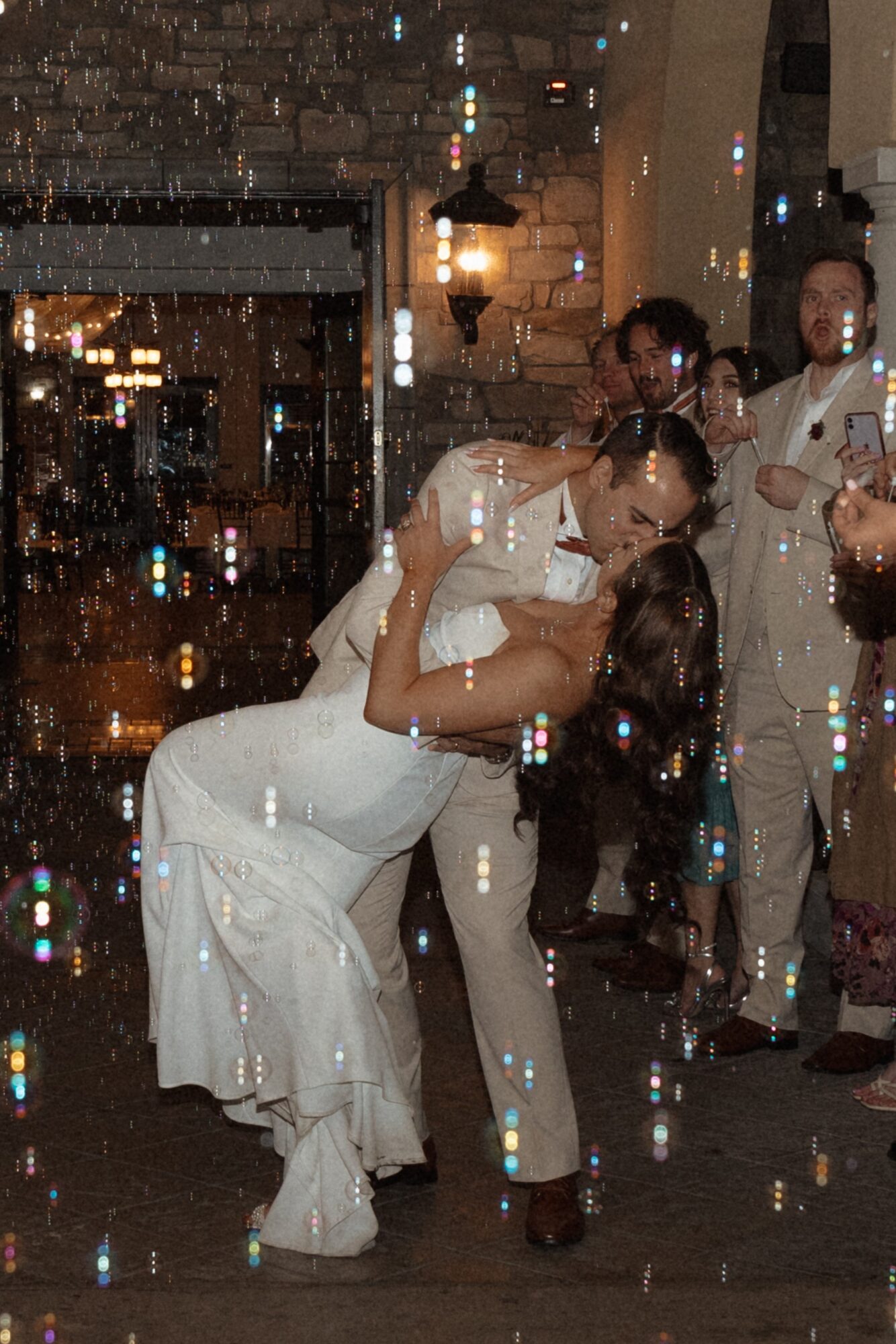 Couple dancing closely at night, surrounded by people, with a brick wall and streetlights in the background.