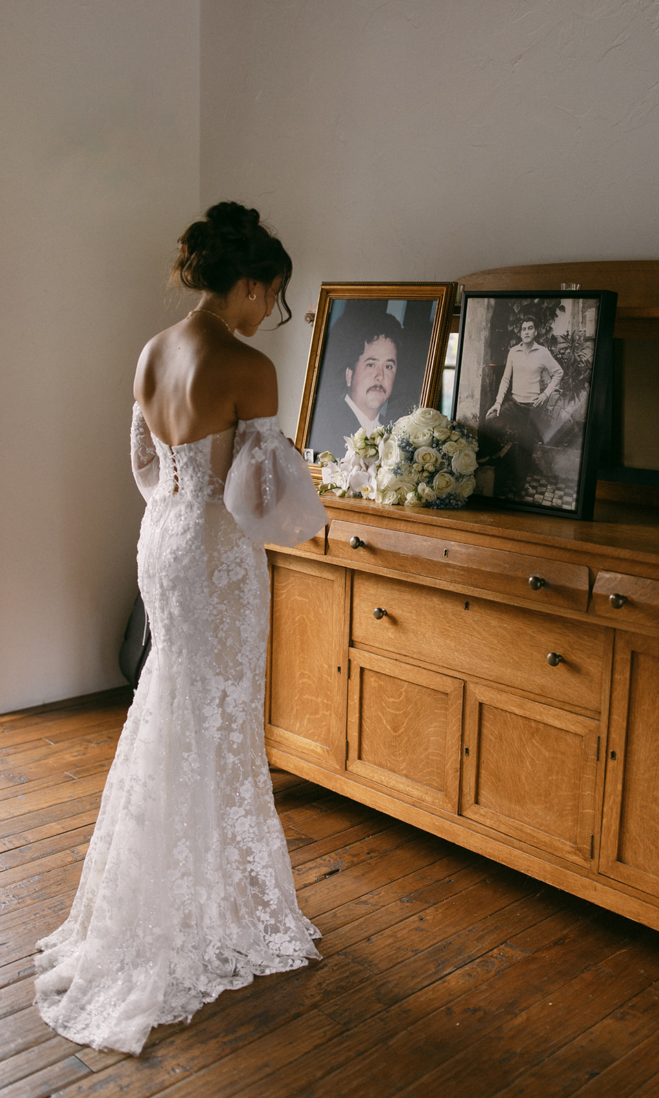 Woman in a white lace dress stands in front of a wooden sideboard with framed photos and flowers, facing away from the camera.