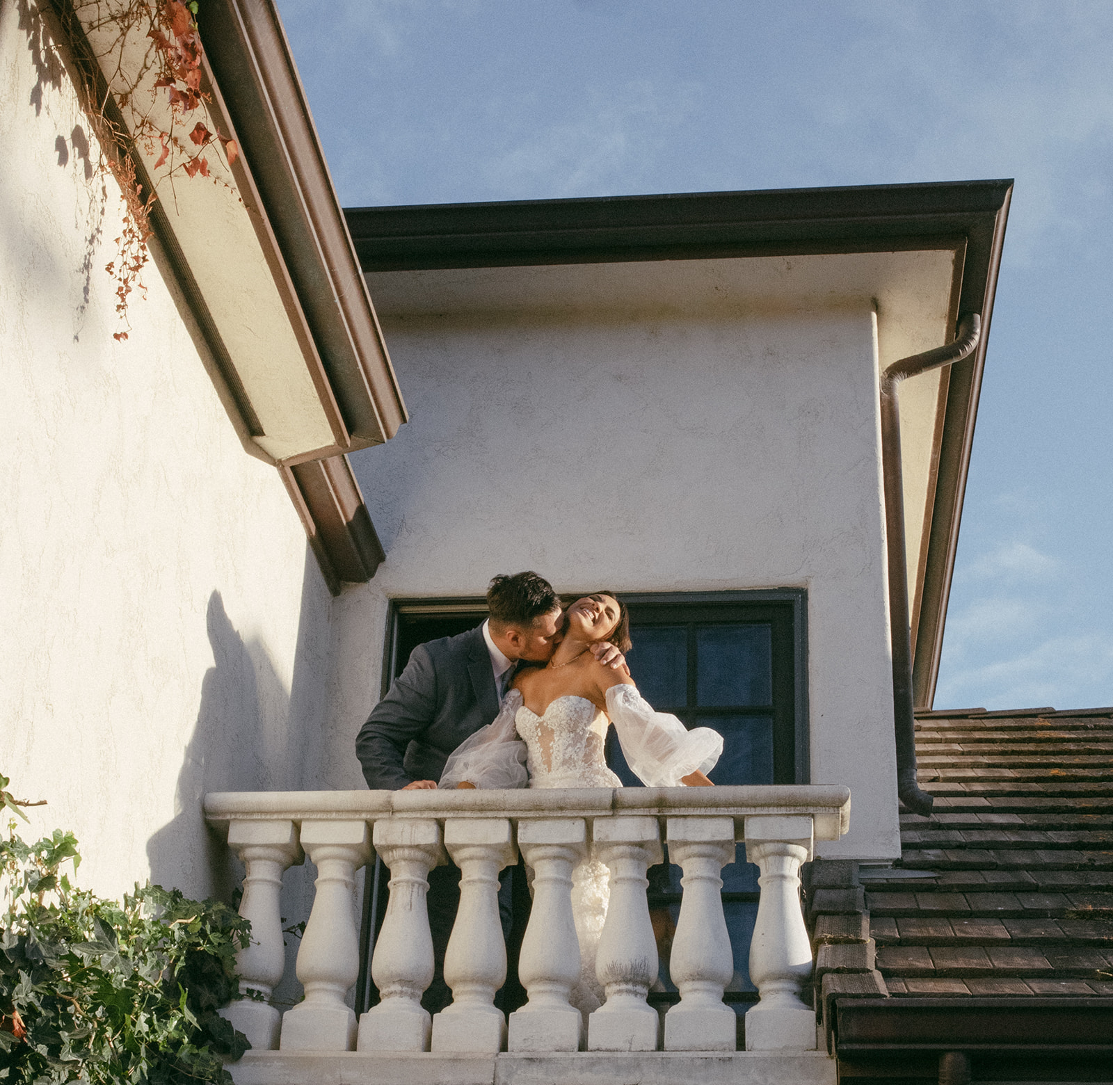 A couple on a balcony, with the man leaning in to kiss the woman, against a white building and blue sky.