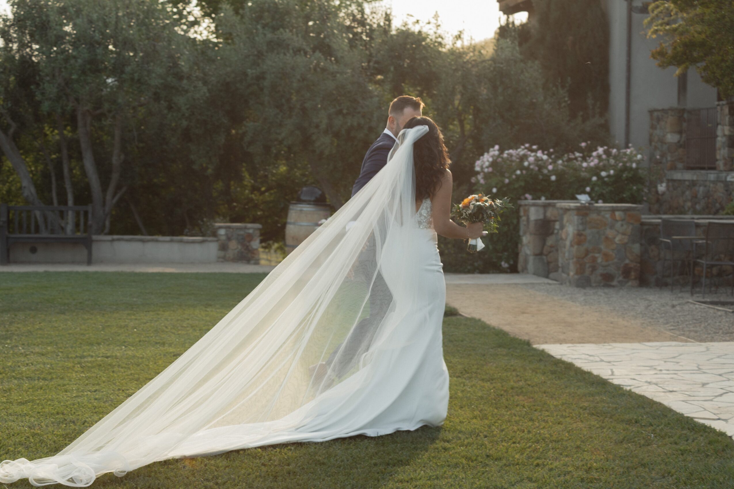A bride and groom stand close together outdoors, with the bride holding a bouquet, and the bride's long veil flowing behind her.
