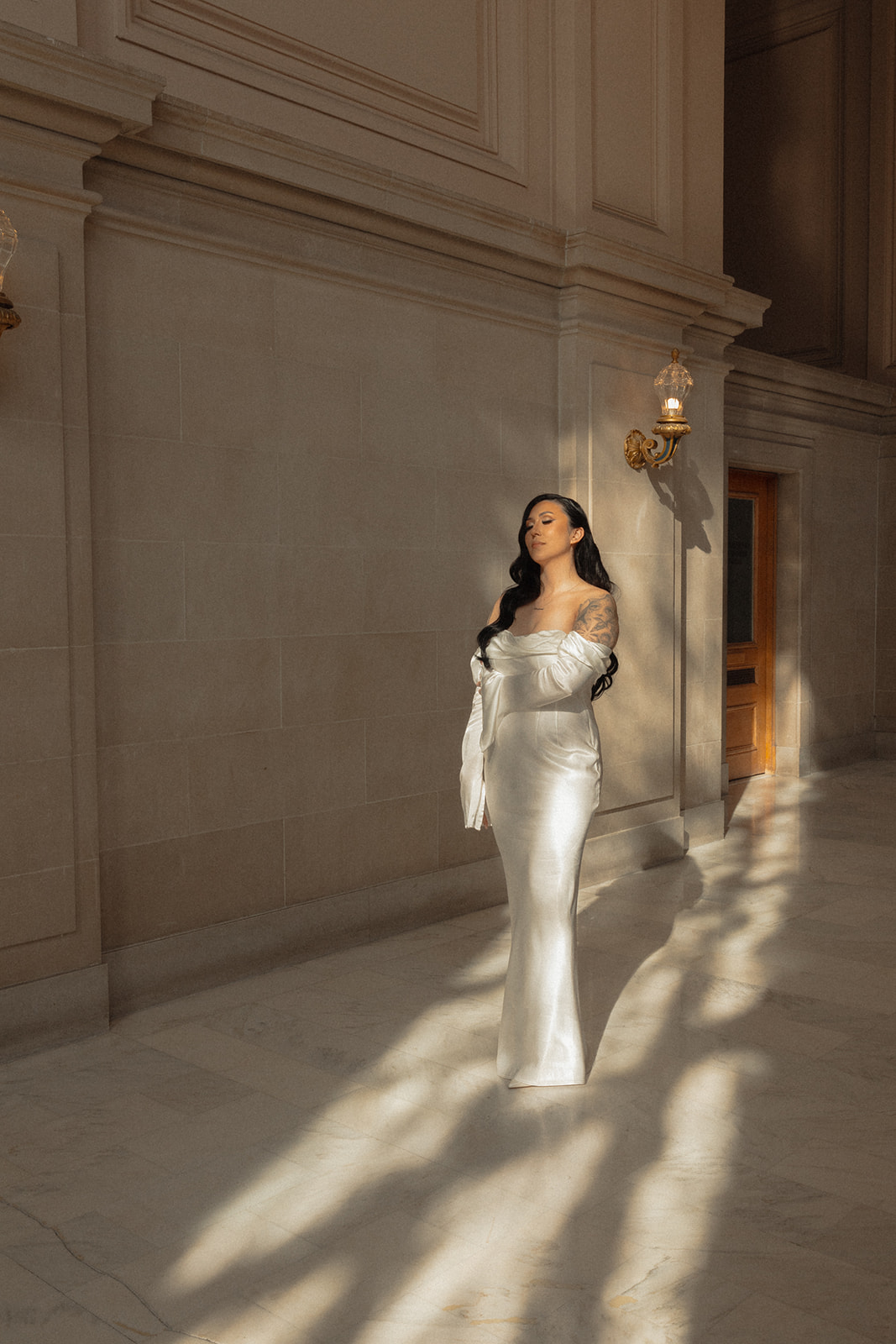 Woman in a white dress walking in a grand hallway with sunlight and shadows on the floor.
