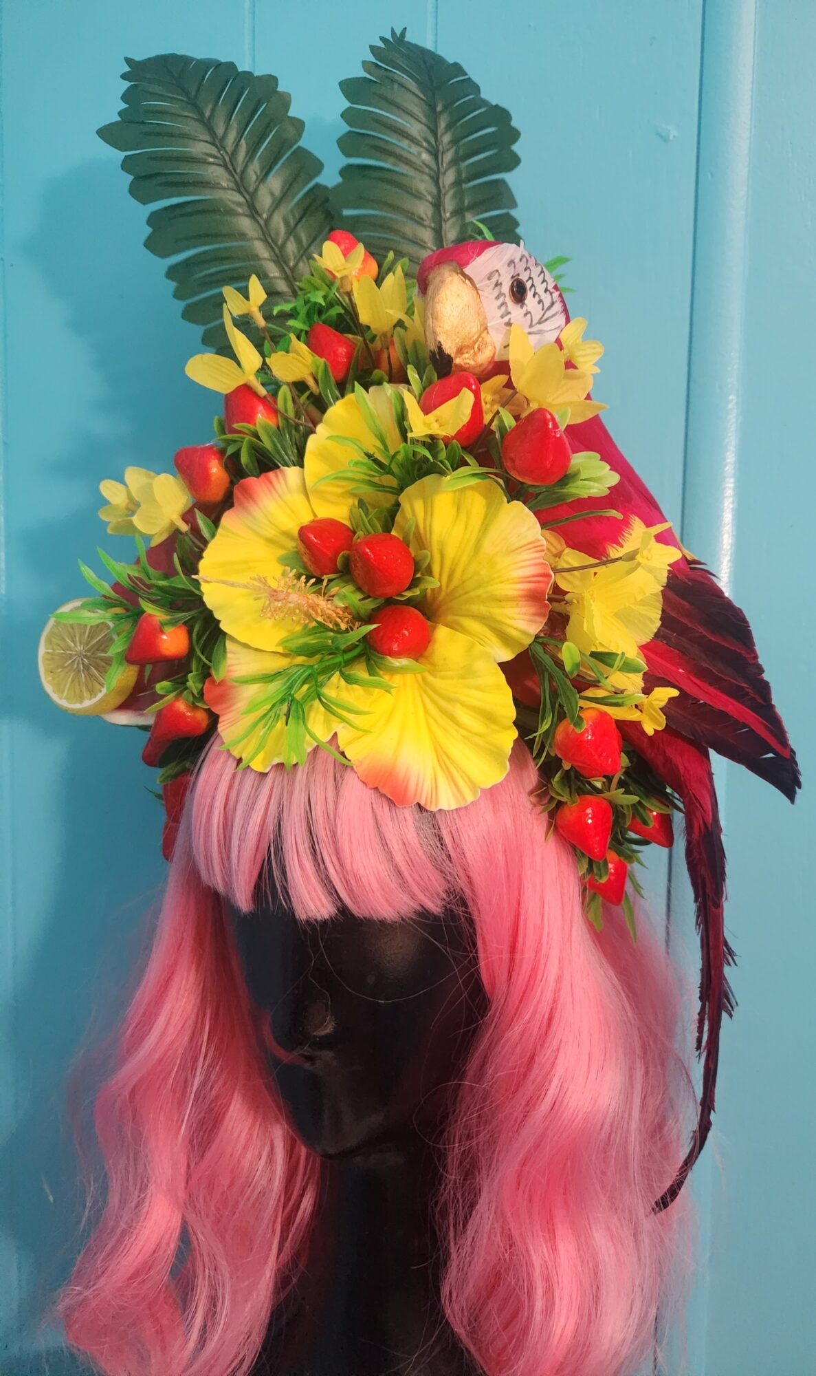 A floral arrangement with yellow, red, and pink flowers, green leaves, and a pink wig on a black mannequin head.