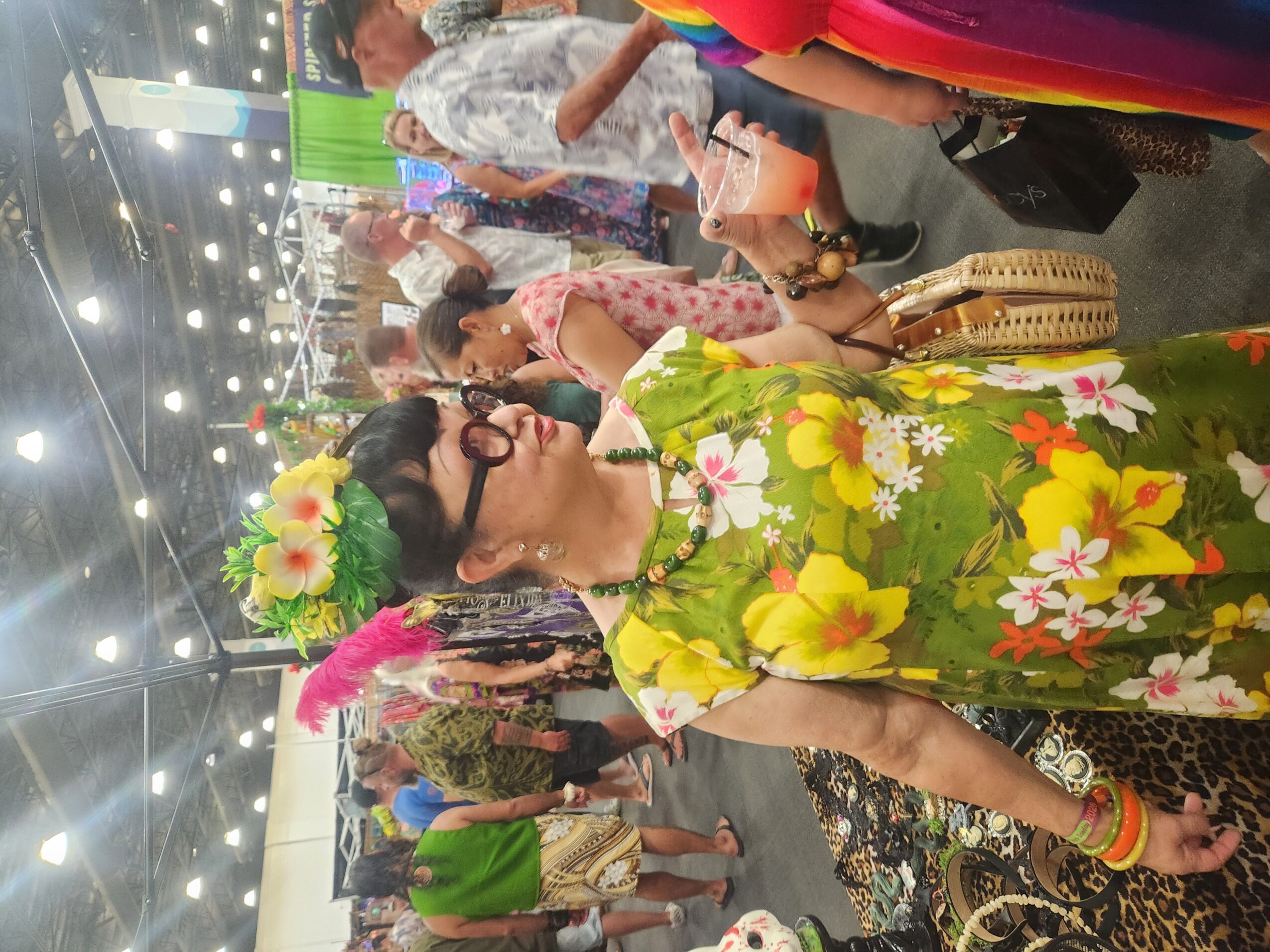 Person wearing a floral dress with a flower crown, sunglasses, and colorful accessories at an indoor event.