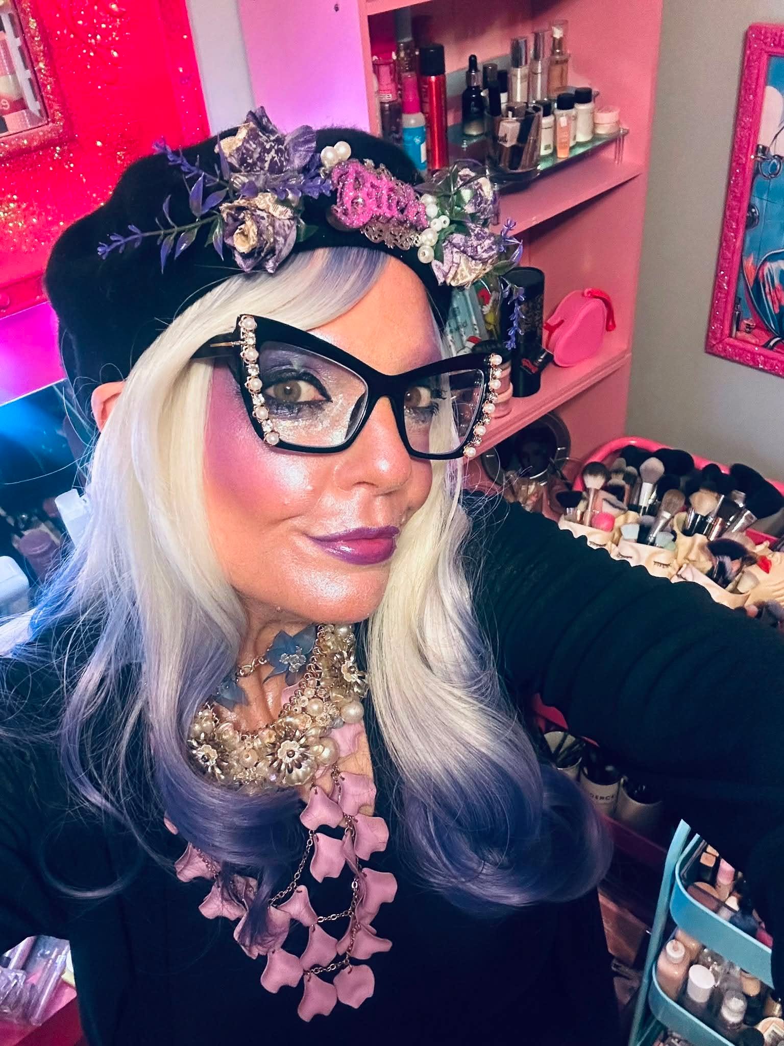 Woman with long, wavy, silver hair wearing glasses and a floral headpiece, in a pink-themed room with makeup products.