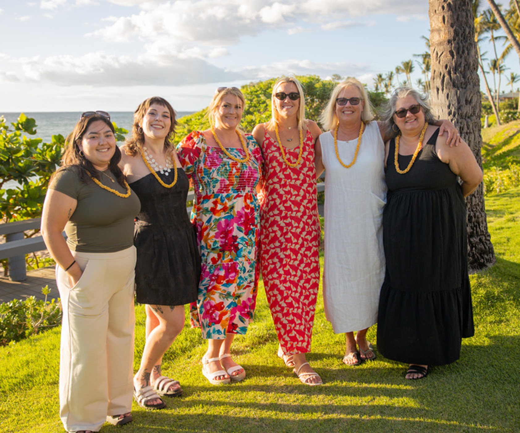 Six women standing outdoors on grass, smiling, with trees and a body of water in the background.