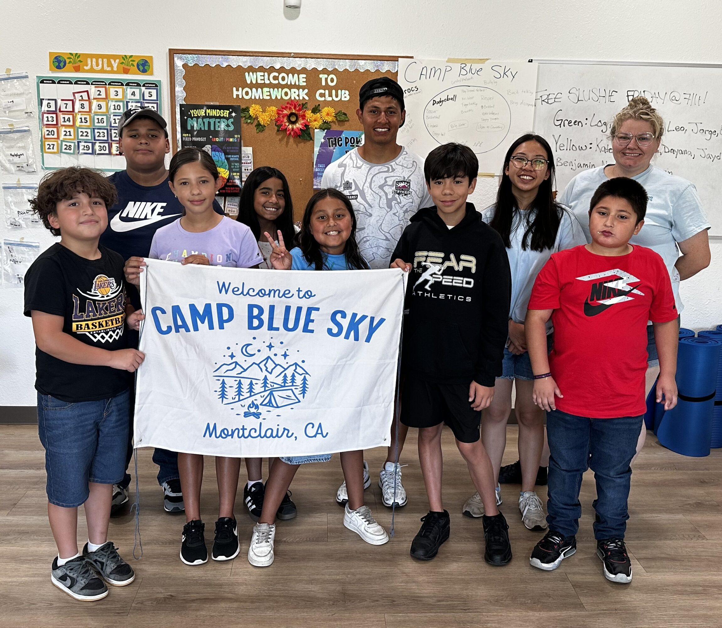 Group of children and an adult holding a banner that reads 'Welcome to Camp Blue Sky, Montclair, CA' in a classroom.