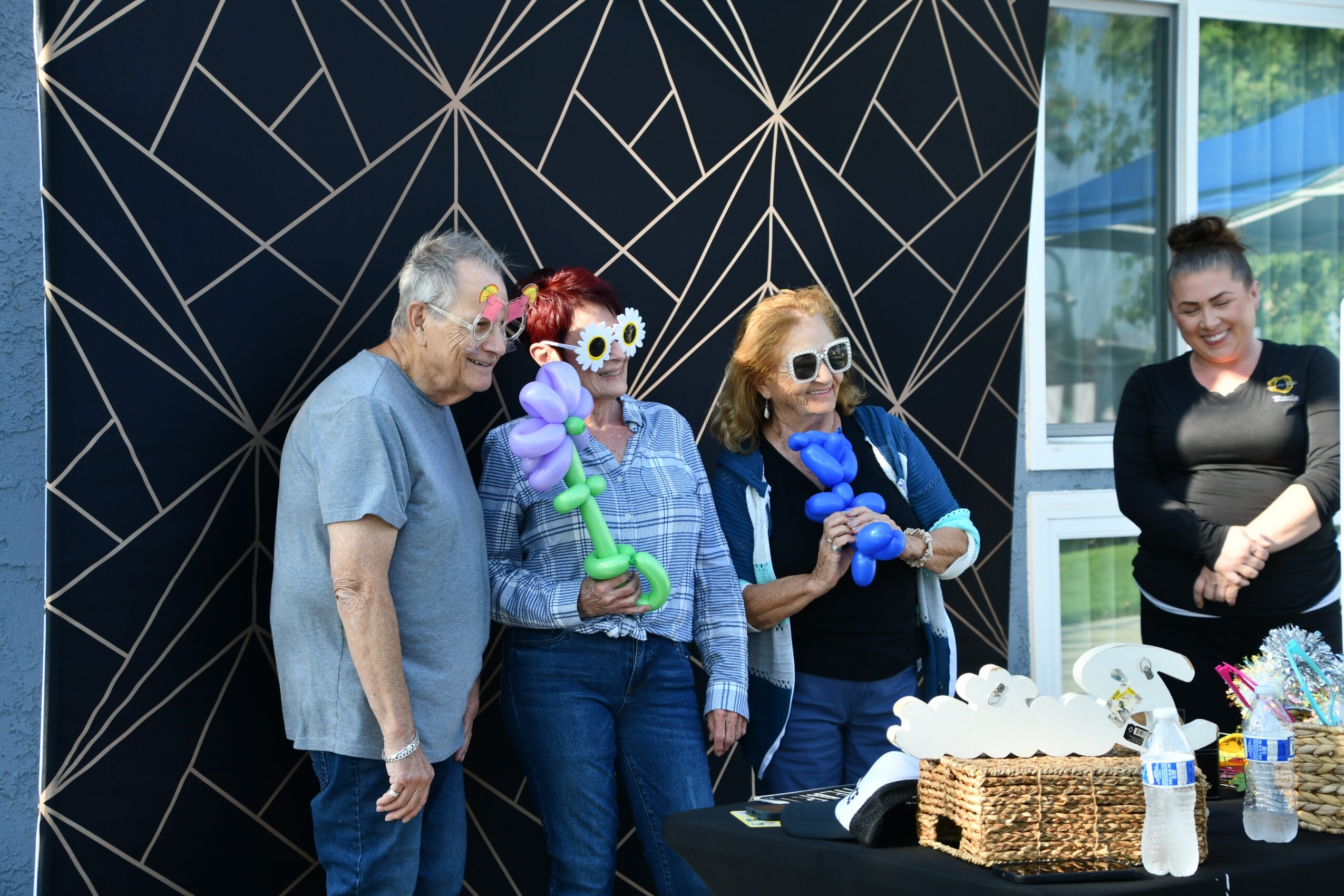 Four women stand together indoors, three wearing sunglasses and holding balloon animals, with a woman on the right smiling, near a window.