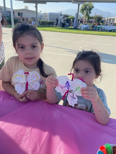 Two young girls sitting at a pink table outdoors, one drinking from a cup, with a park and buildings in background.
