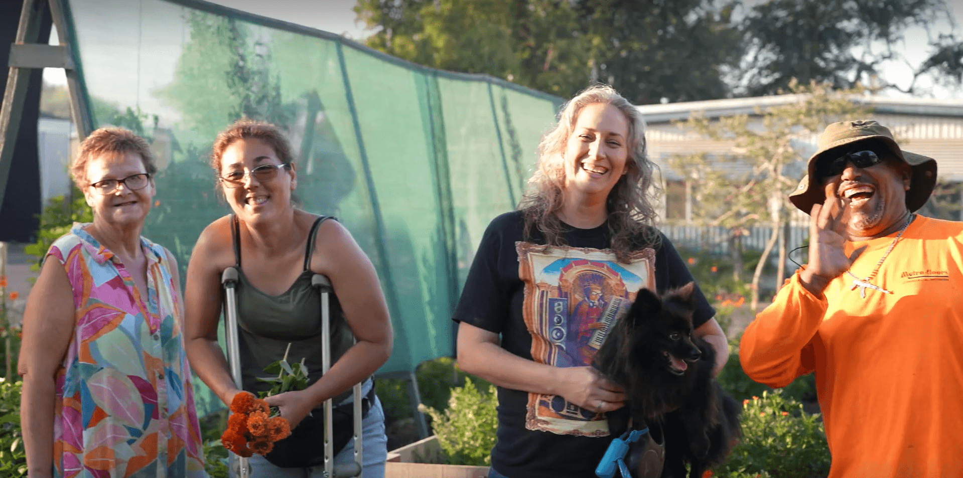 Four women standing outdoors, smiling, with trees and a glass building in the background.