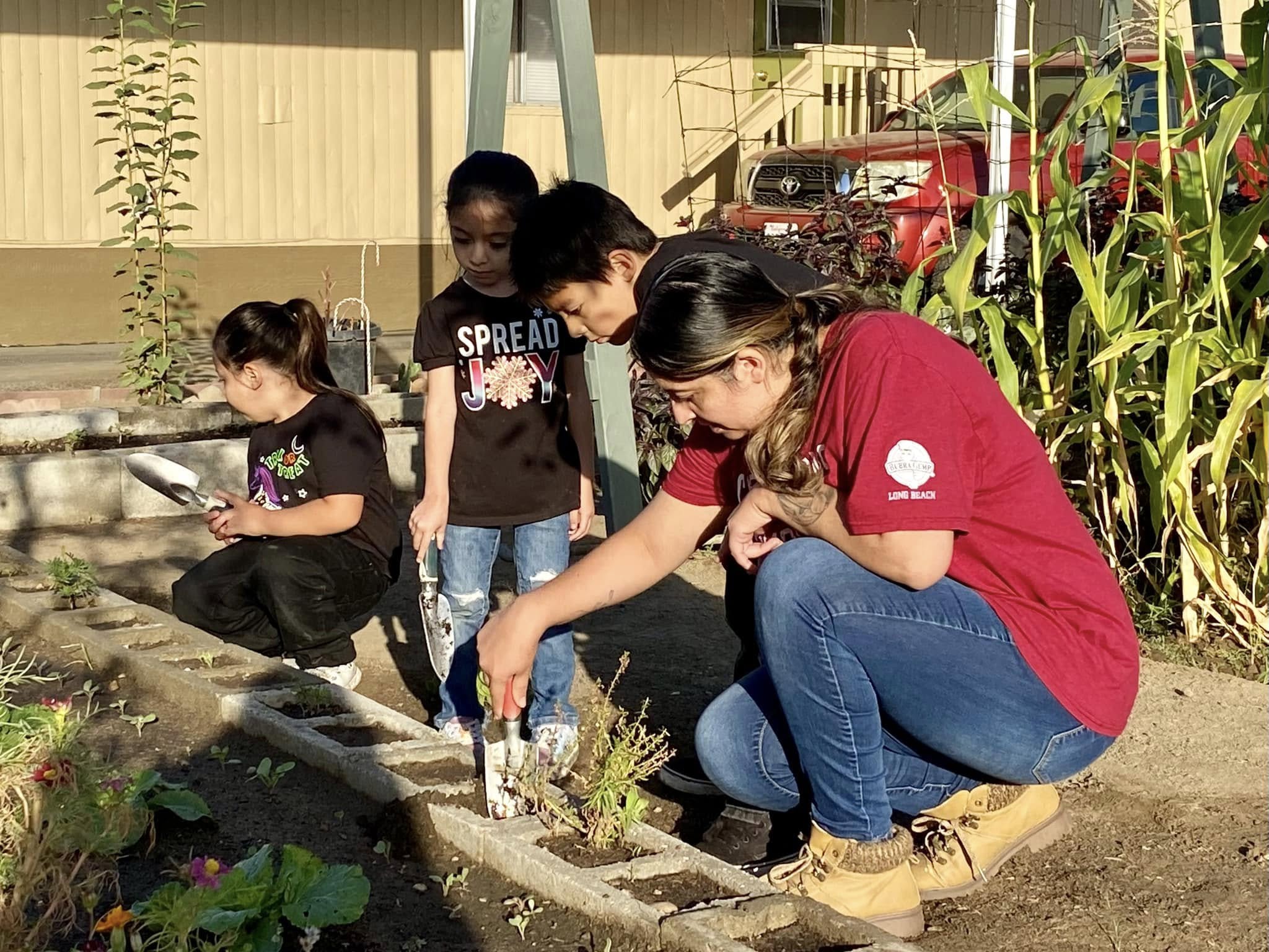 Four people, including children and an adult, planting or tending to a garden outdoors near plants and a building.