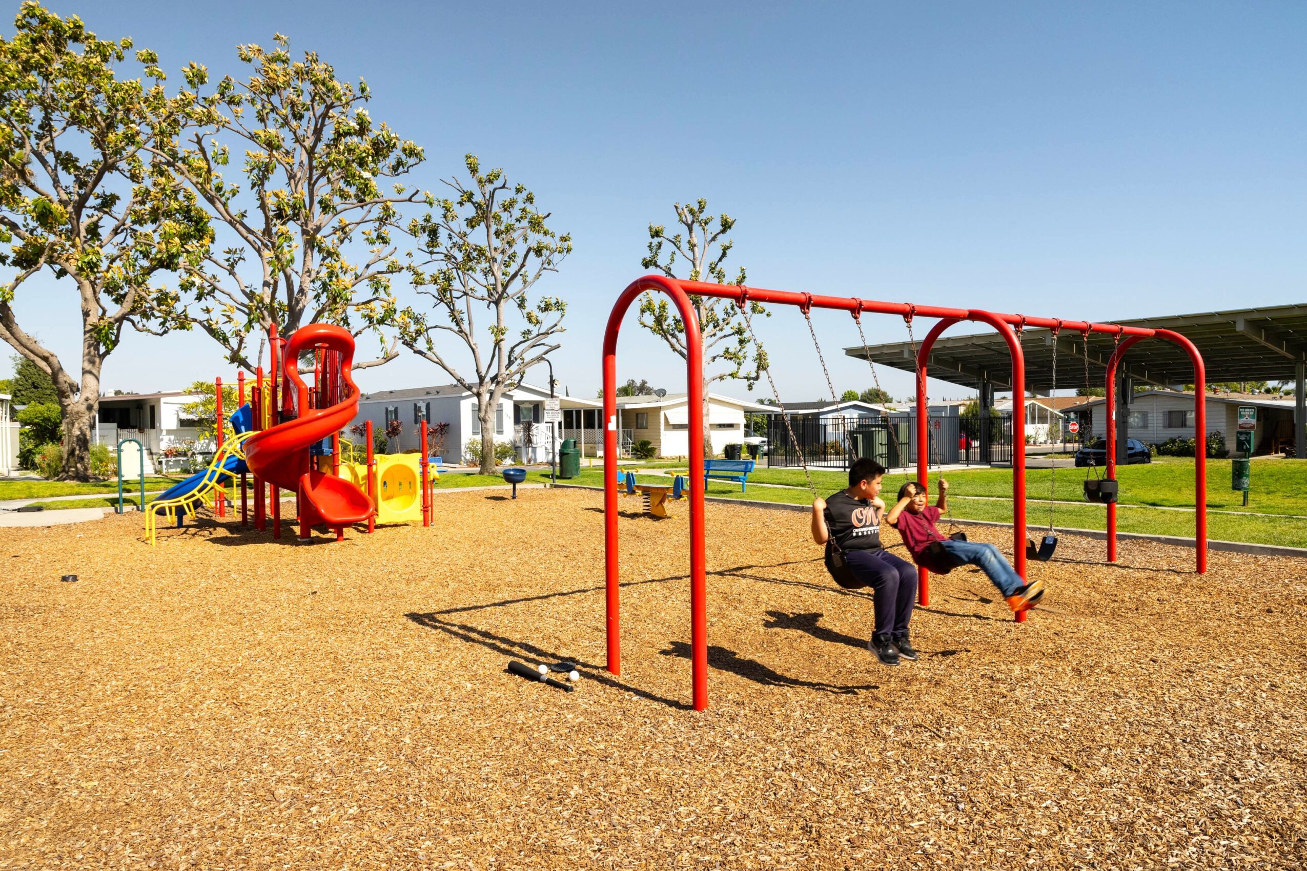 Children playing on swings and slide in a park with trees and a clear sky.