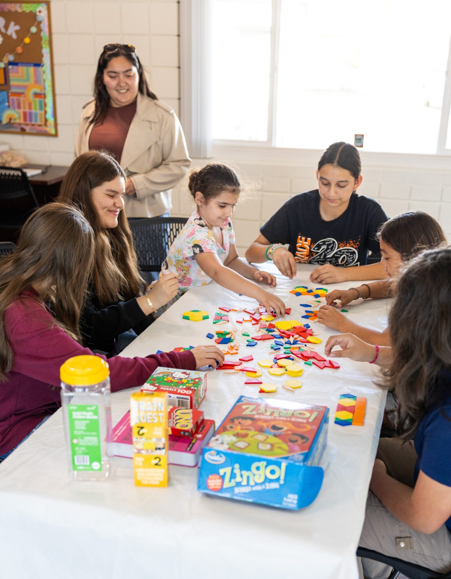 Group of children playing a board game at a table with a woman supervising, in a bright room with large windows.