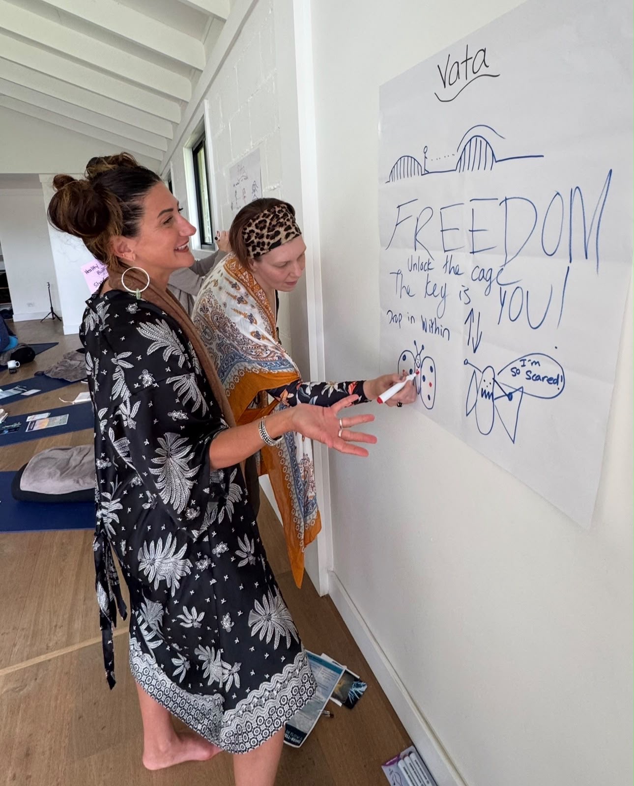 Two women writing on a large paper on the wall, one smiling, in a bright room with white walls and windows.