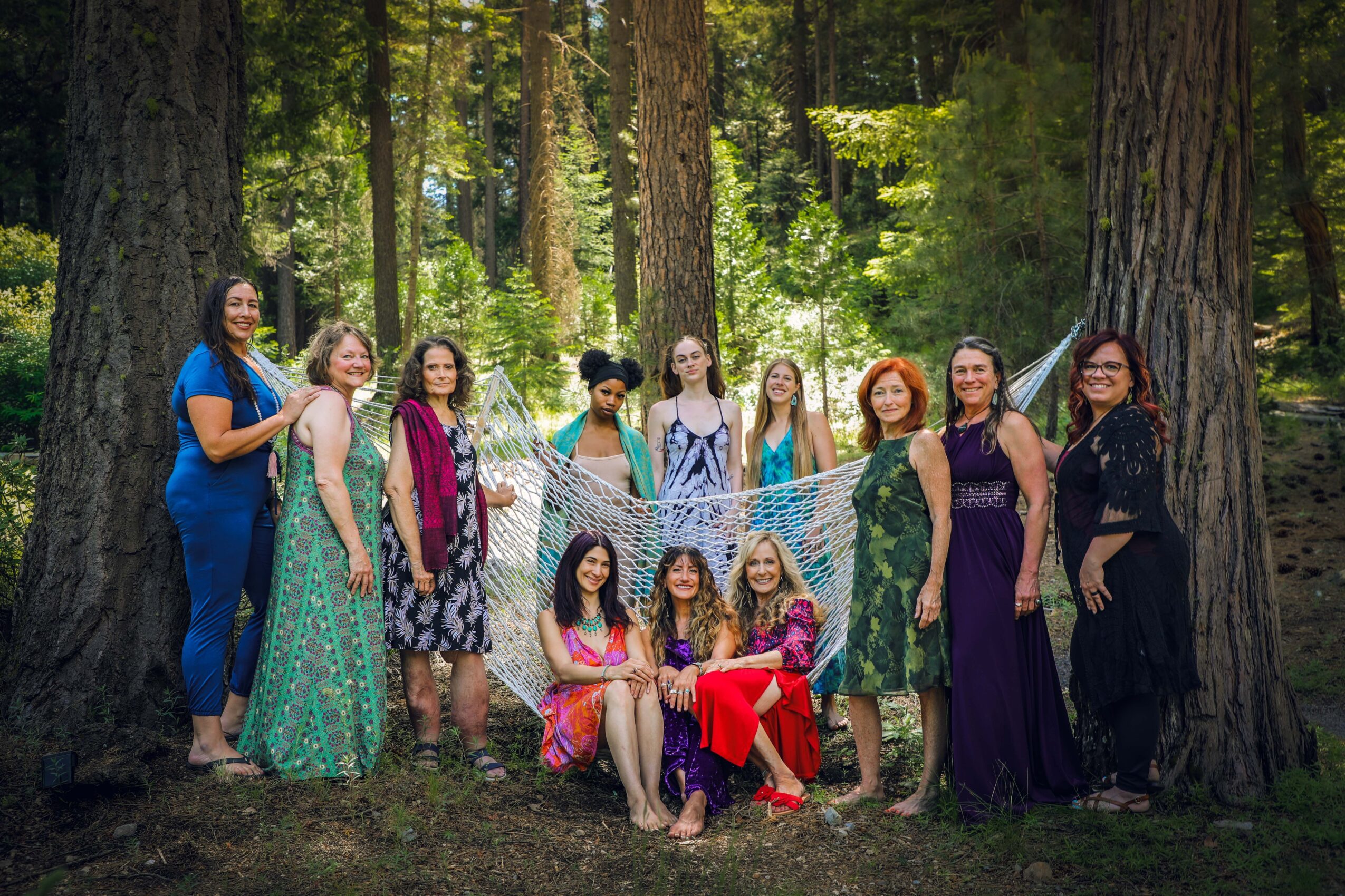 Group of ten women standing and sitting in a forest, some holding a net, surrounded by tall trees and greenery.