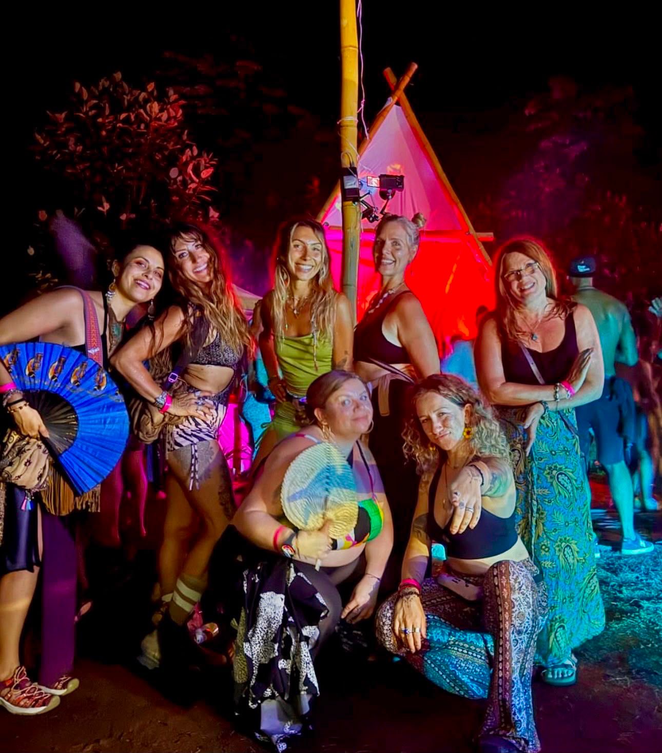 Group of women in colorful costumes at night with a tent and trees in background.