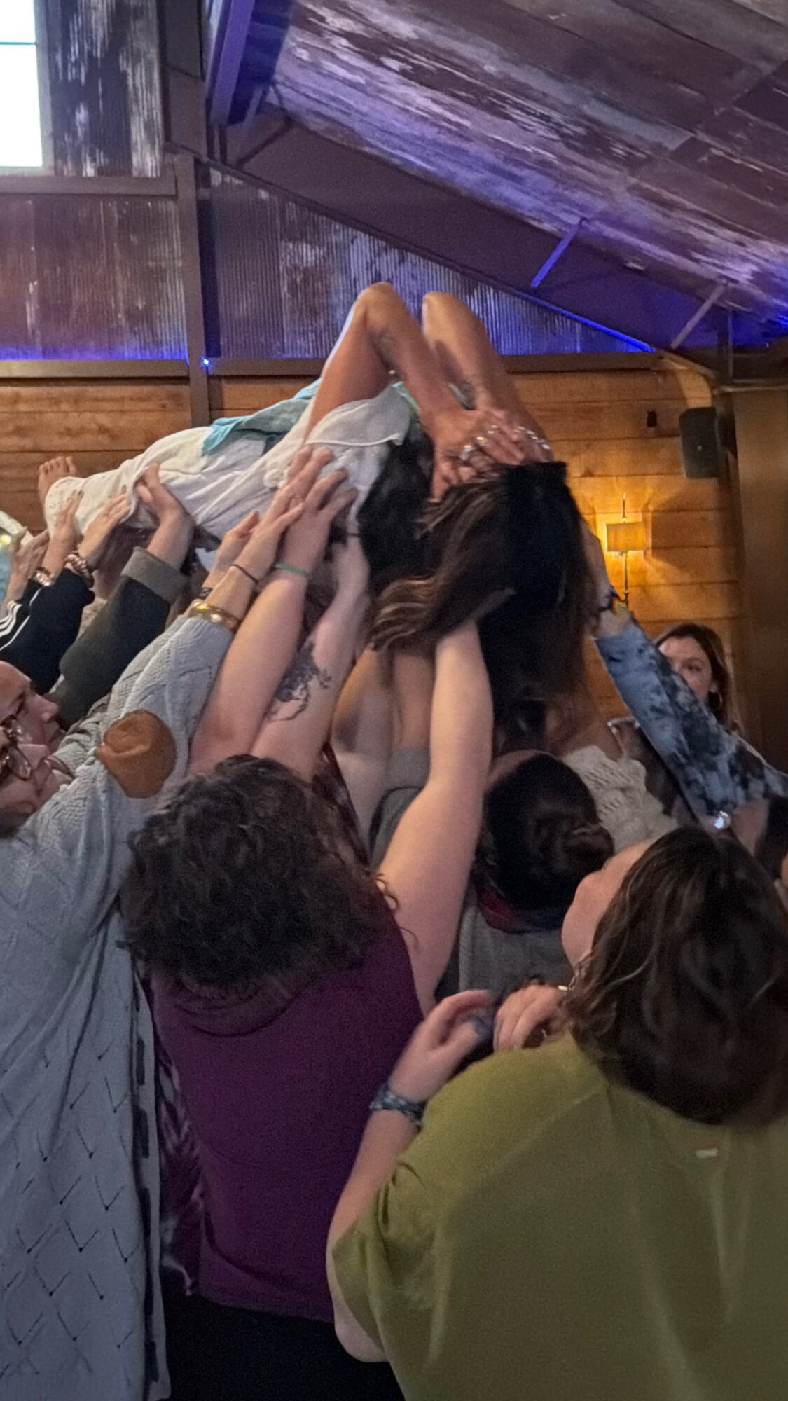 Group of people lifting a person in a crowd, indoors with wooden walls and ceiling, some reaching out.