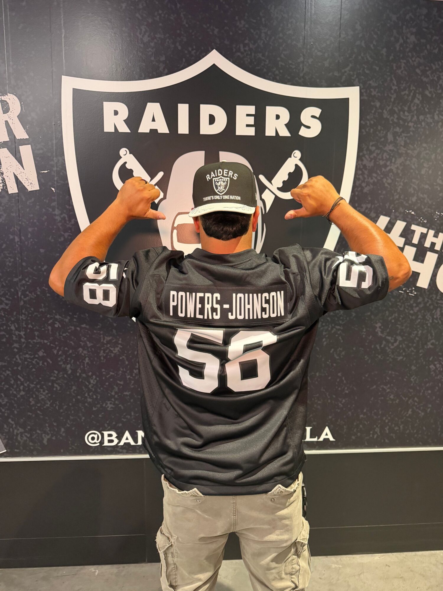 Person wearing Raiders gear, including a cap and jersey, standing in front of Raiders logo background, giving thumbs up.