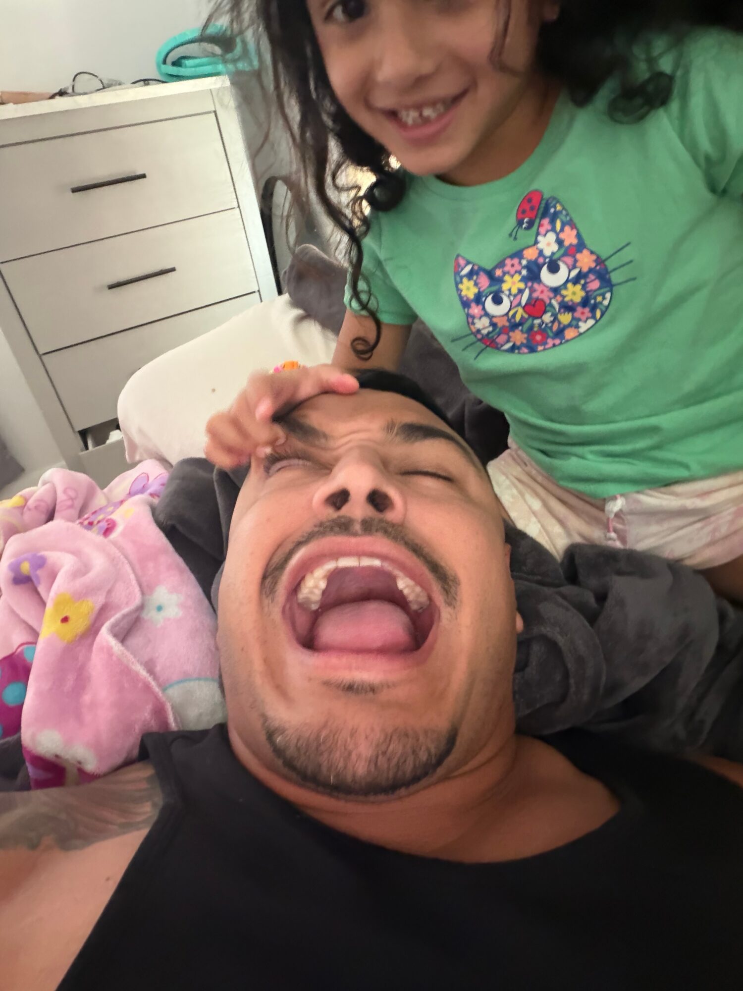 Child smiling and leaning over a man lying down, who is making a funny face with his mouth open.
