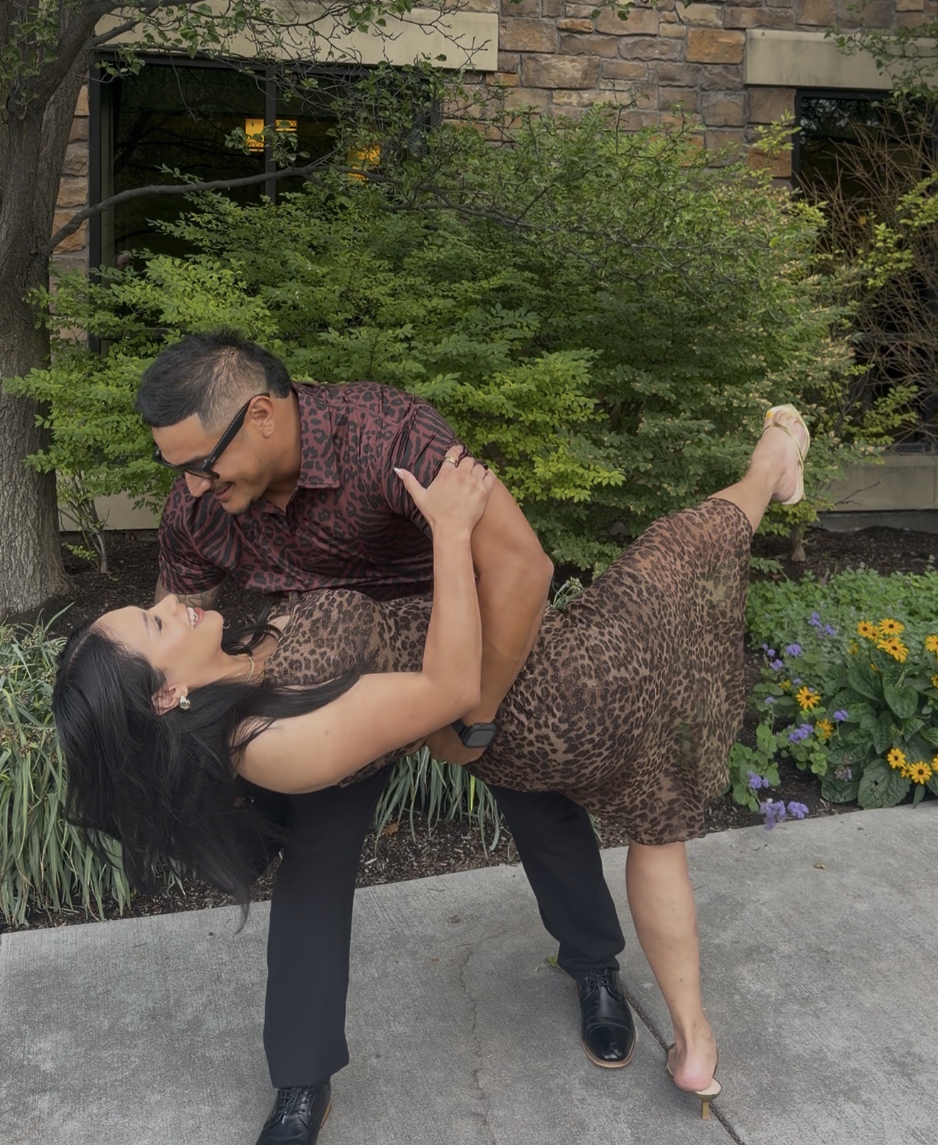 A man and woman dance outdoors on a sidewalk with greenery and flowers in the background.