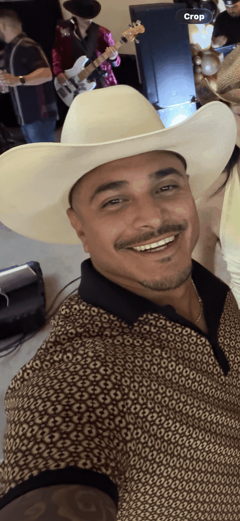 Man wearing a large white hat and patterned shirt, smiling at the camera, with a band playing in the background.