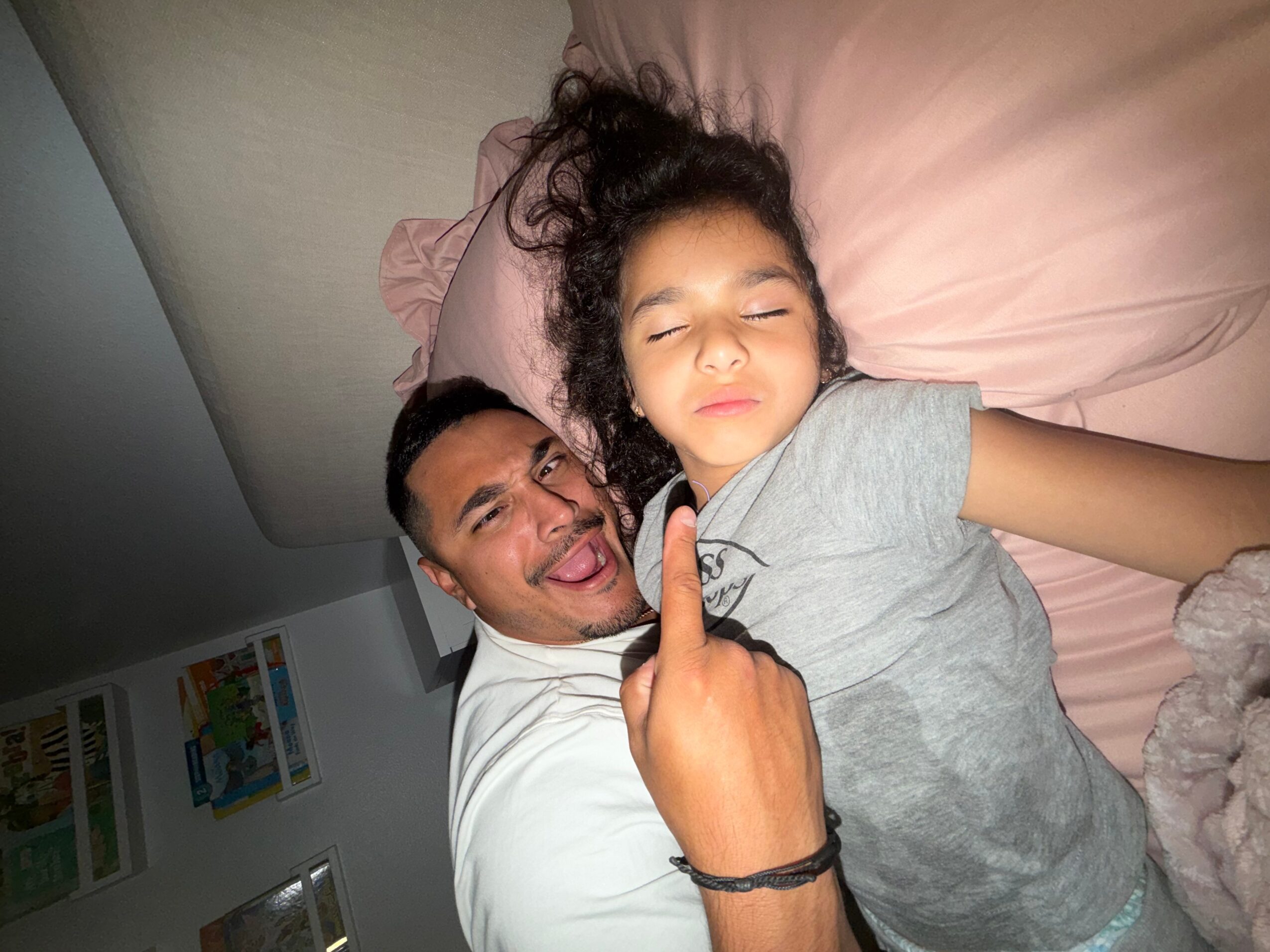 Adult and child lying in bed, smiling and making a peace sign, with a wall and pictures in the background.