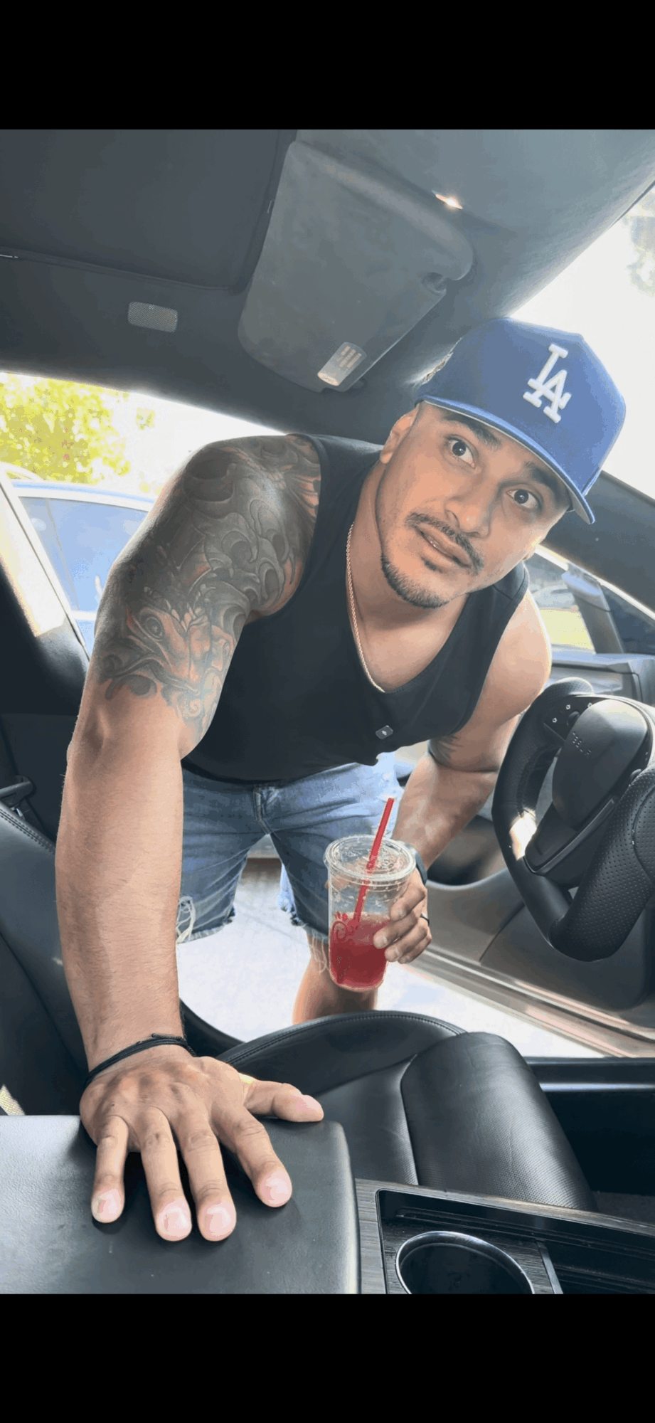 Man leaning into car, wearing a blue cap, black tank top, holding a drink with a straw, inside vehicle.