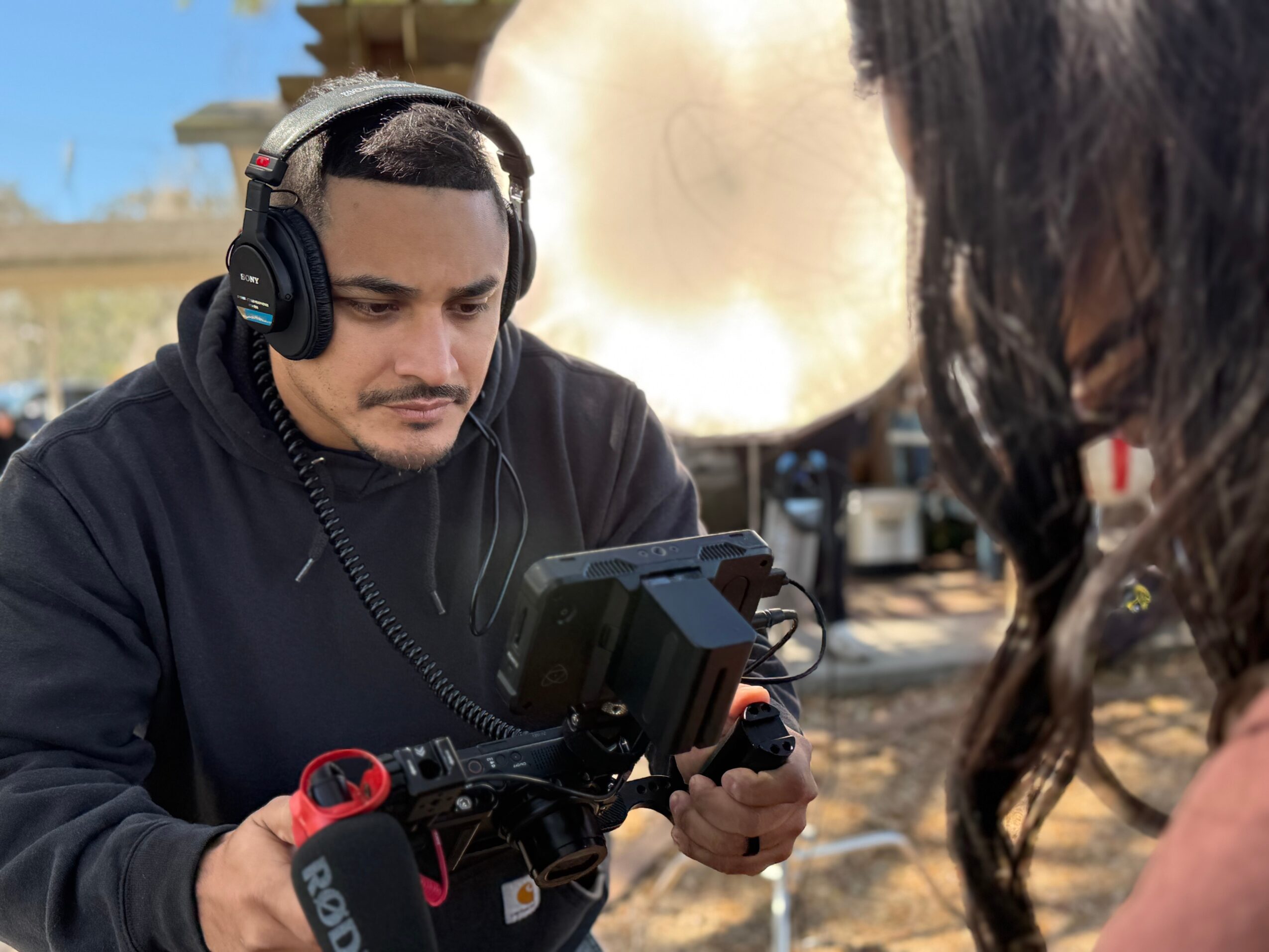 Man wearing headphones looks at a monitor, with a person partially visible nearby, outdoors with equipment in background.