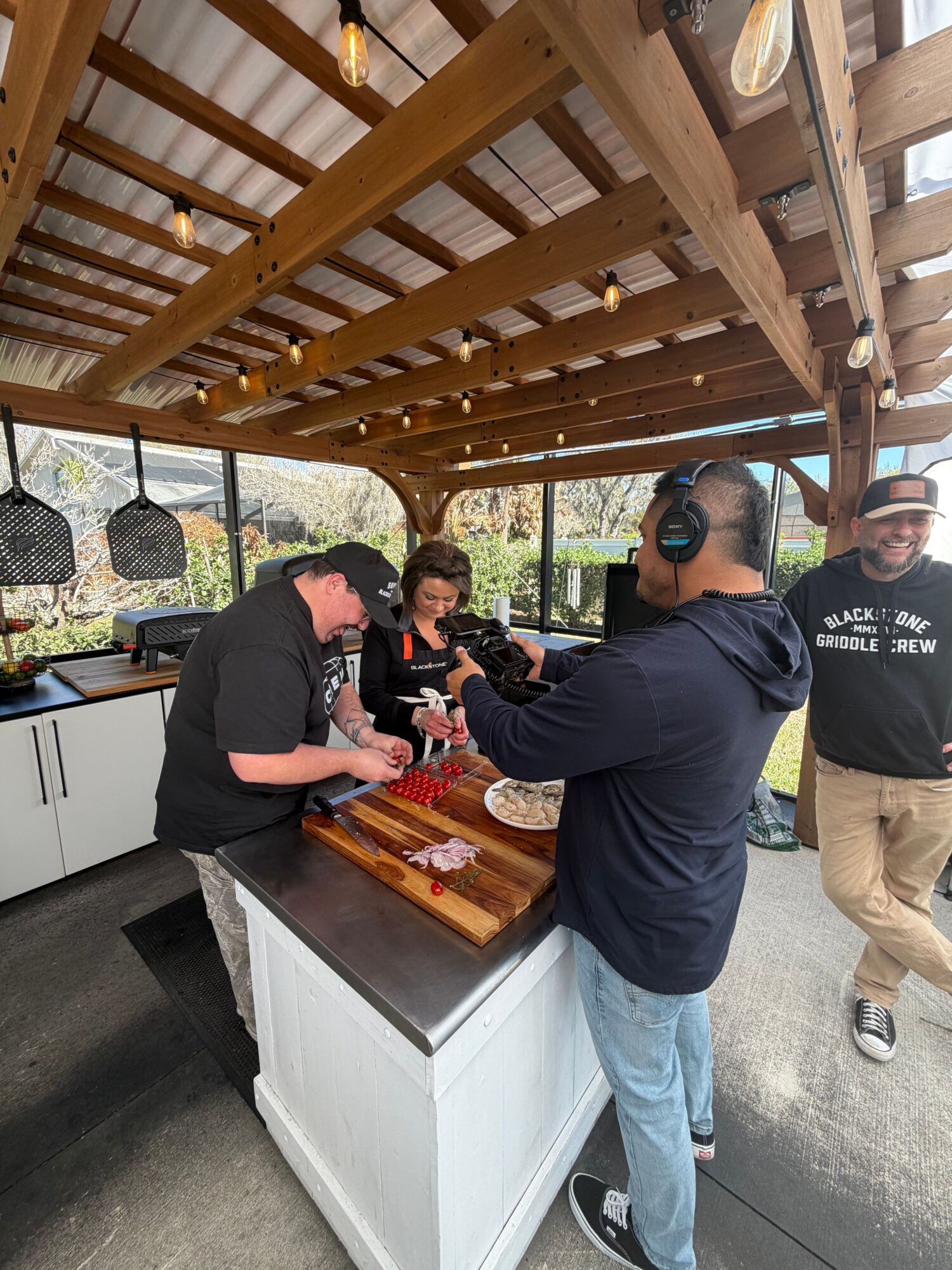 Two men are preparing food at an outdoor wooden bar with a person in the background. One man wears headphones and a hoodie.