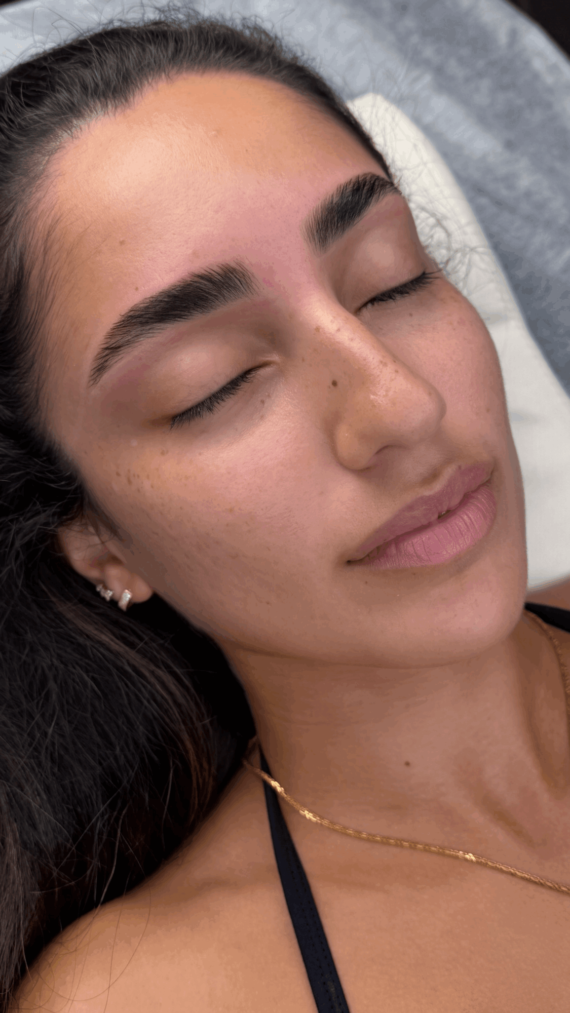 Close-up of a woman with closed eyes, dark hair, and a gold necklace, lying down with a relaxed expression.