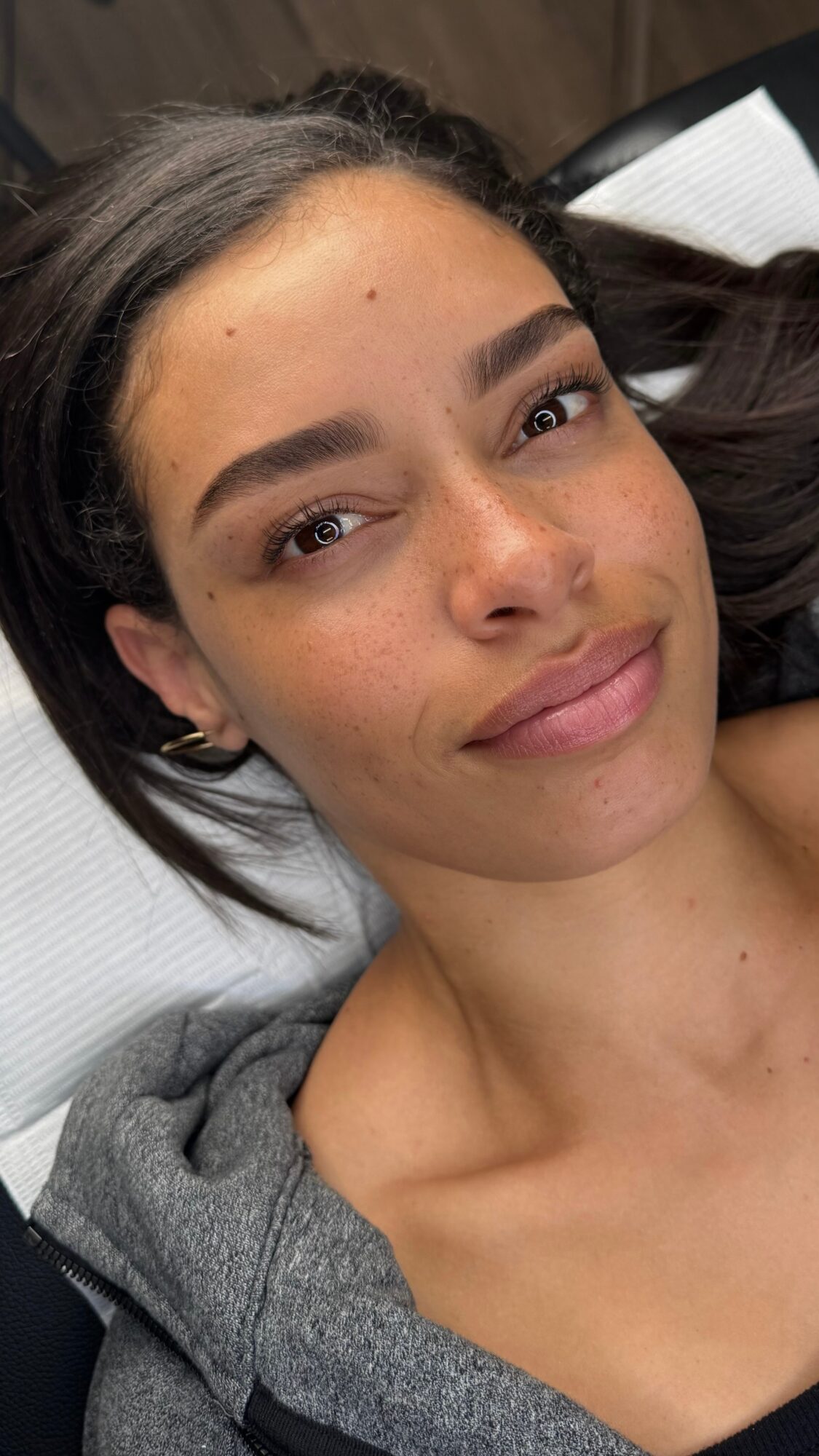 Close-up of a young woman with dark hair, lying on a pillow, smiling softly, wearing a gray top.