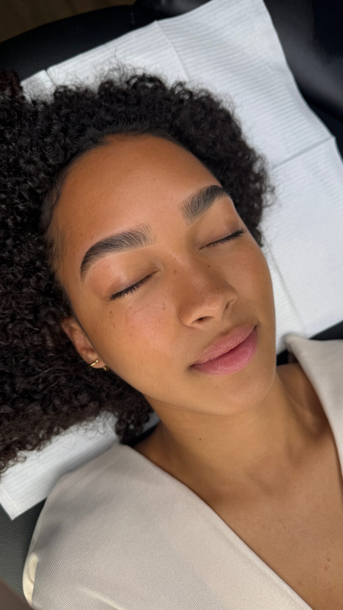 Young woman with curly hair lying with eyes closed on a white pillow, relaxed expression, wearing a light-colored top.