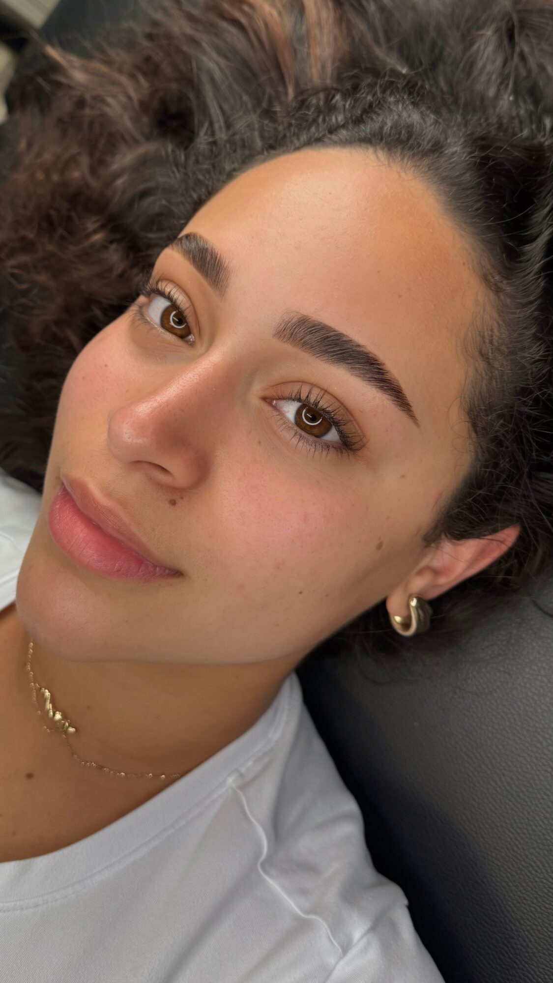 Close-up of a woman with dark, curly hair, light skin, and minimal makeup, lying down and looking at the camera.