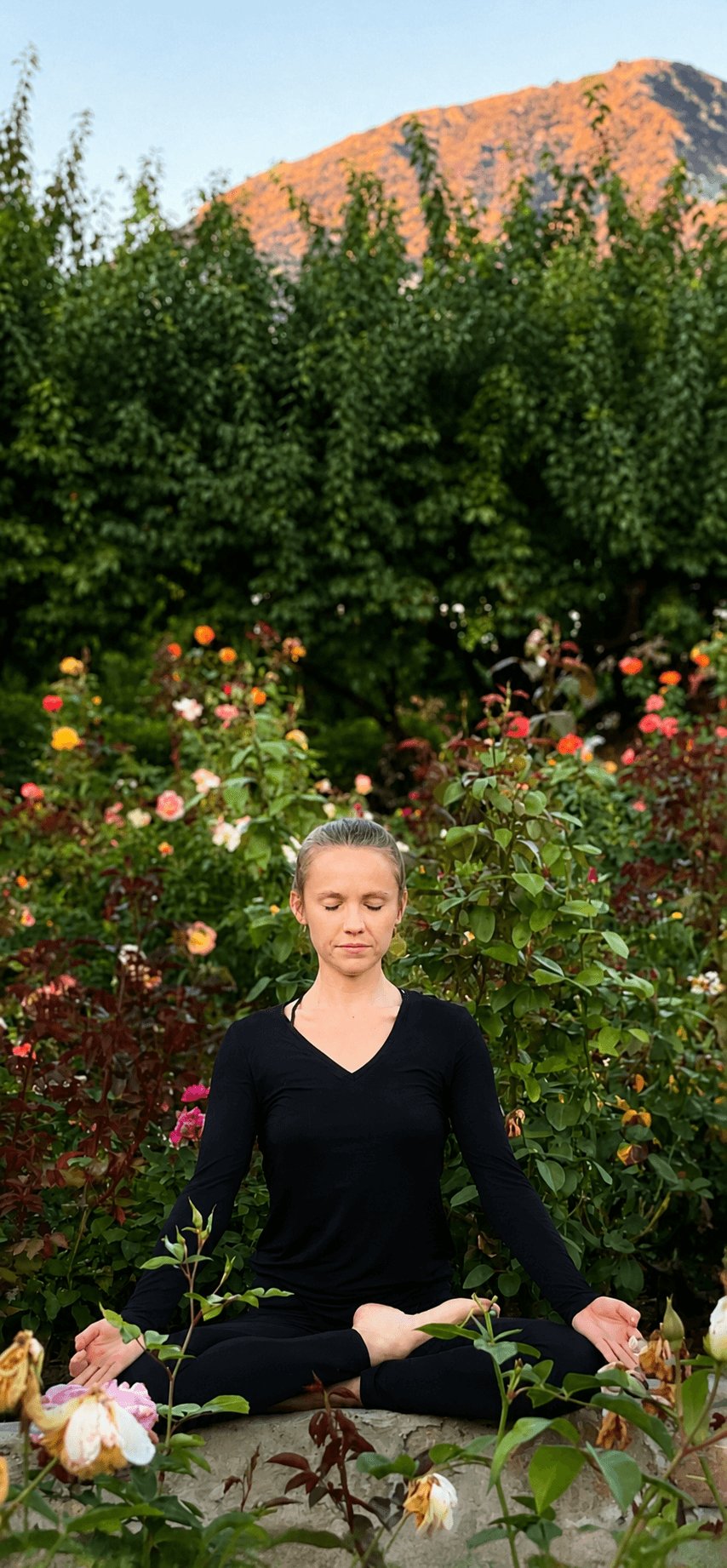 Person practicing yoga in a garden with colorful flowers and green trees, mountain in background, sitting cross-legged with eyes closed.