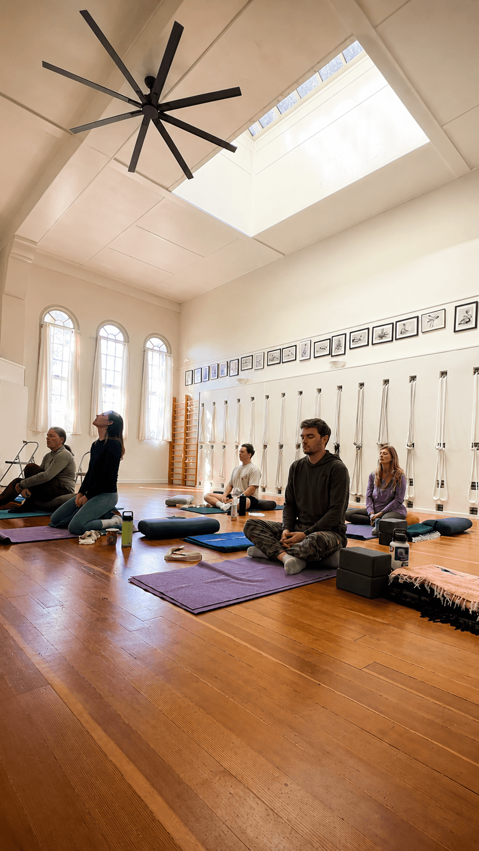 People practicing yoga or meditation on mats in a bright room with large windows and framed pictures on the wall.