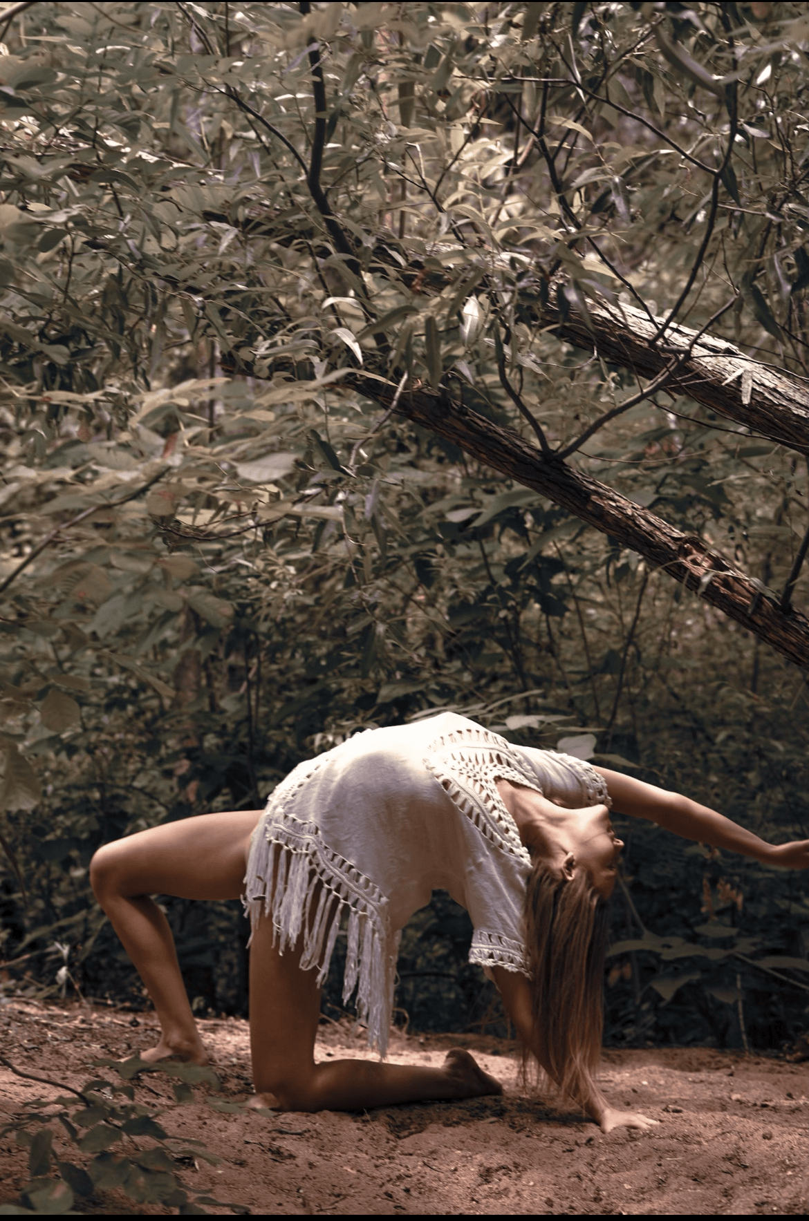 Person in a white dress performing a backbend on the ground in a forest setting.