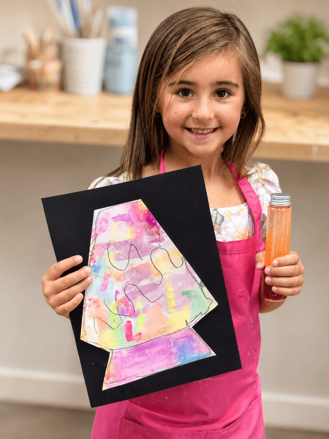 Young girl with long hair smiling, holding a colorful drawing and a bottle of orange liquid, in a room with a wooden shelf.