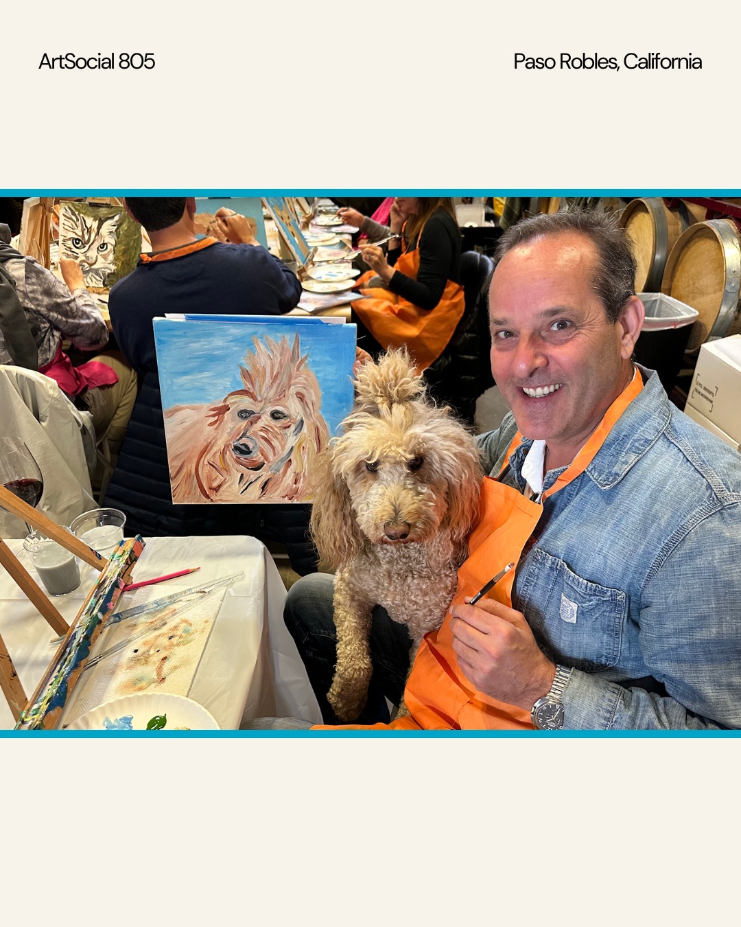 Man holding a dog at an art event with a painting of a dog in the background, people seated at tables, art supplies visible.