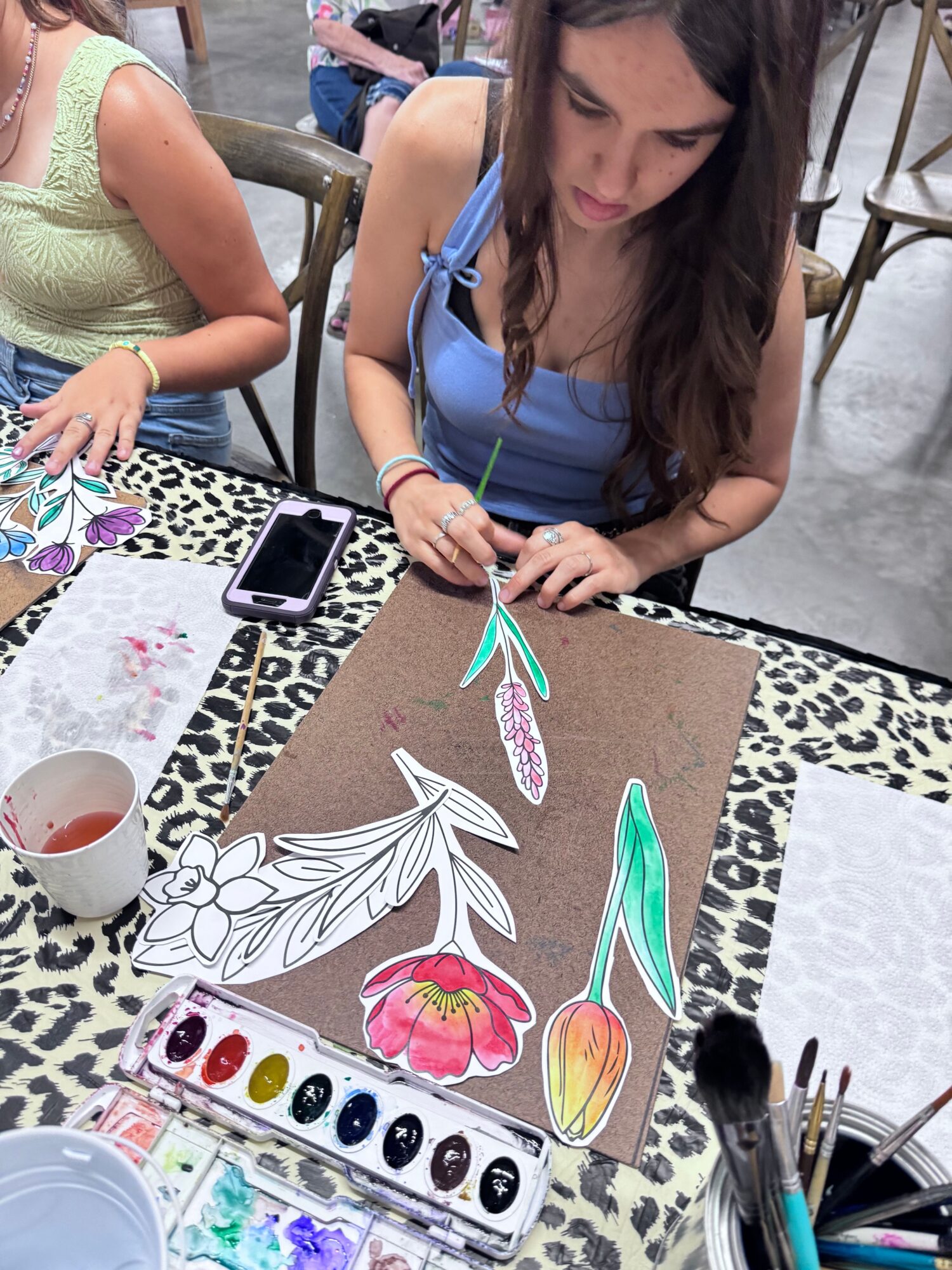 Woman drawing colorful flower and leaf illustrations on paper with watercolor paints and brushes, sitting at a table.