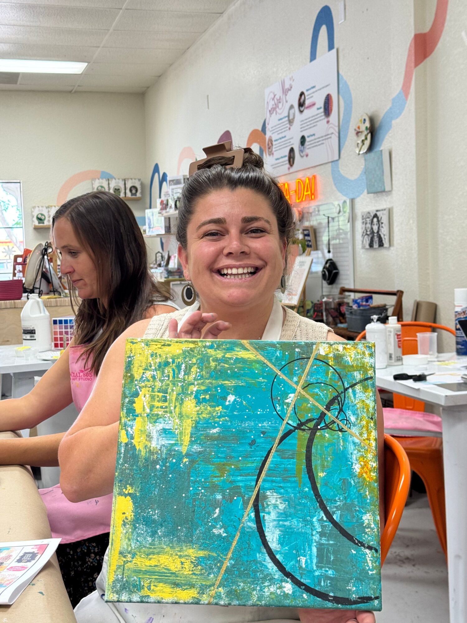Two women smiling behind a colorful abstract painting on a table in a room with art supplies and posters.