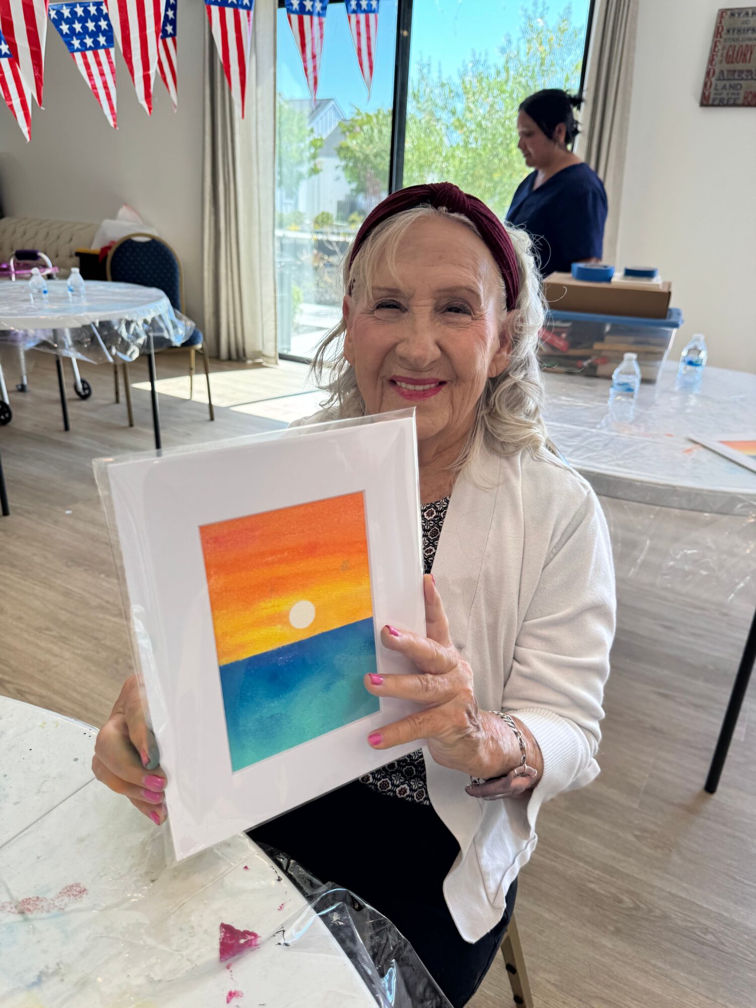 Woman holding colorful sunset artwork in a room with American flags and people in the background.