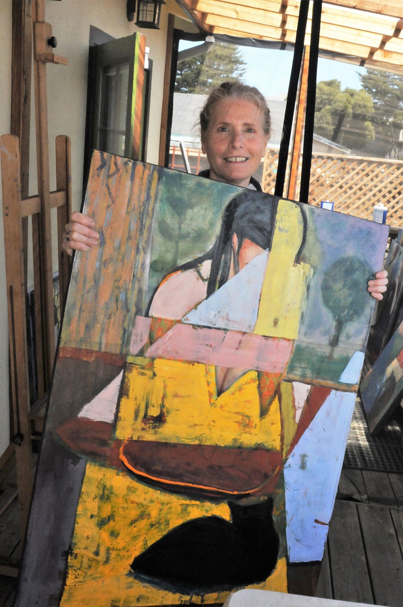 Child holding a large abstract painting with colorful shapes and figures, smiling indoors near windows.