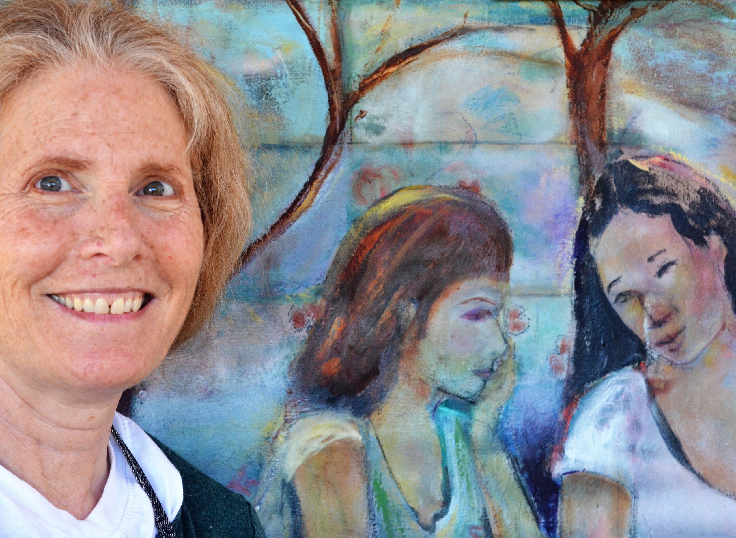 Woman with red hair smiling in front of a colorful painting of two girls, one with brown hair and one with black hair.