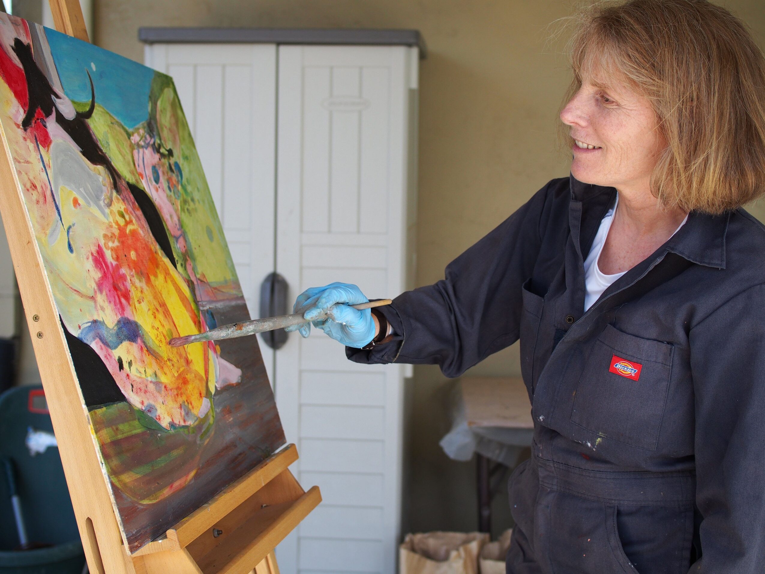 Woman painting on canvas with colorful abstract artwork, smiling, wearing a dark jacket and blue gloves, in a room with a white cabinet.