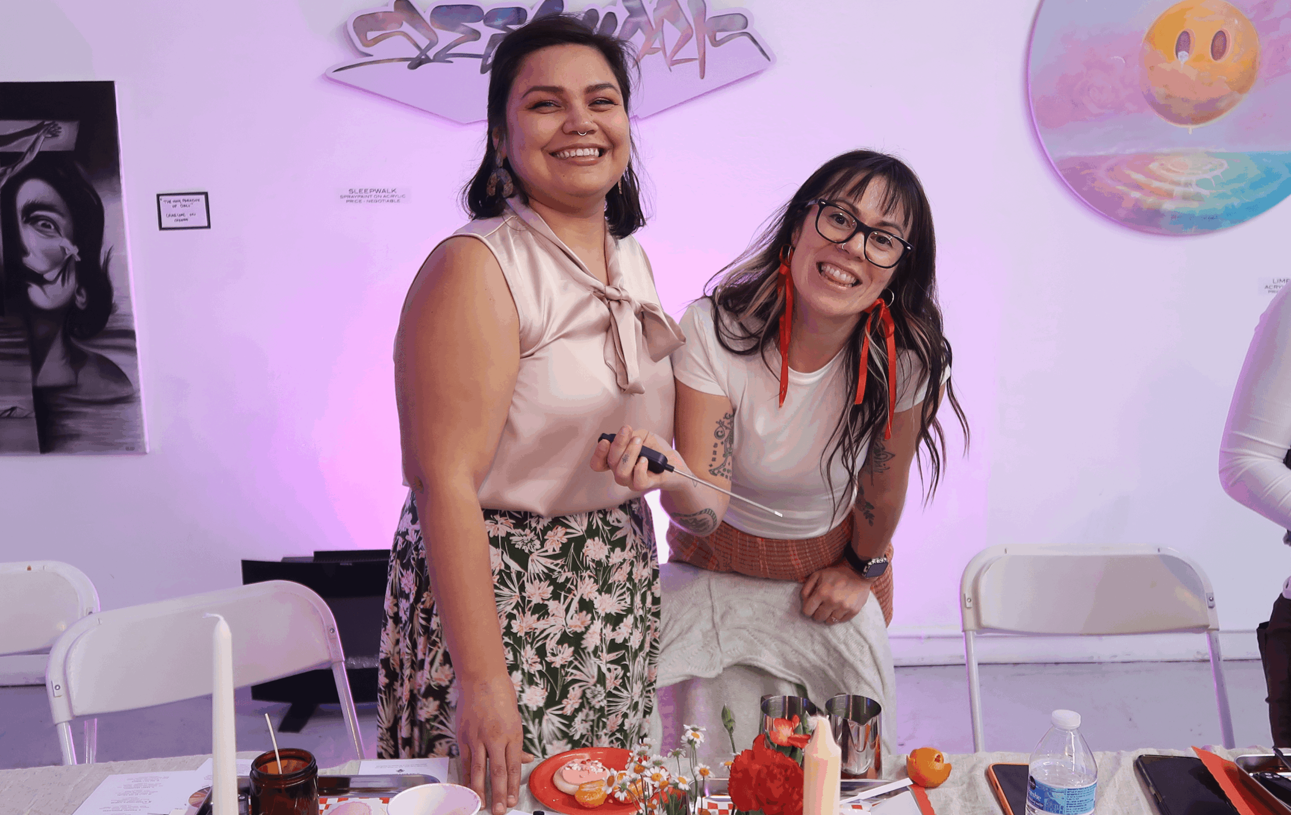 Two women smiling and holding hands at a celebration table with cake and decorations, in a brightly lit room.