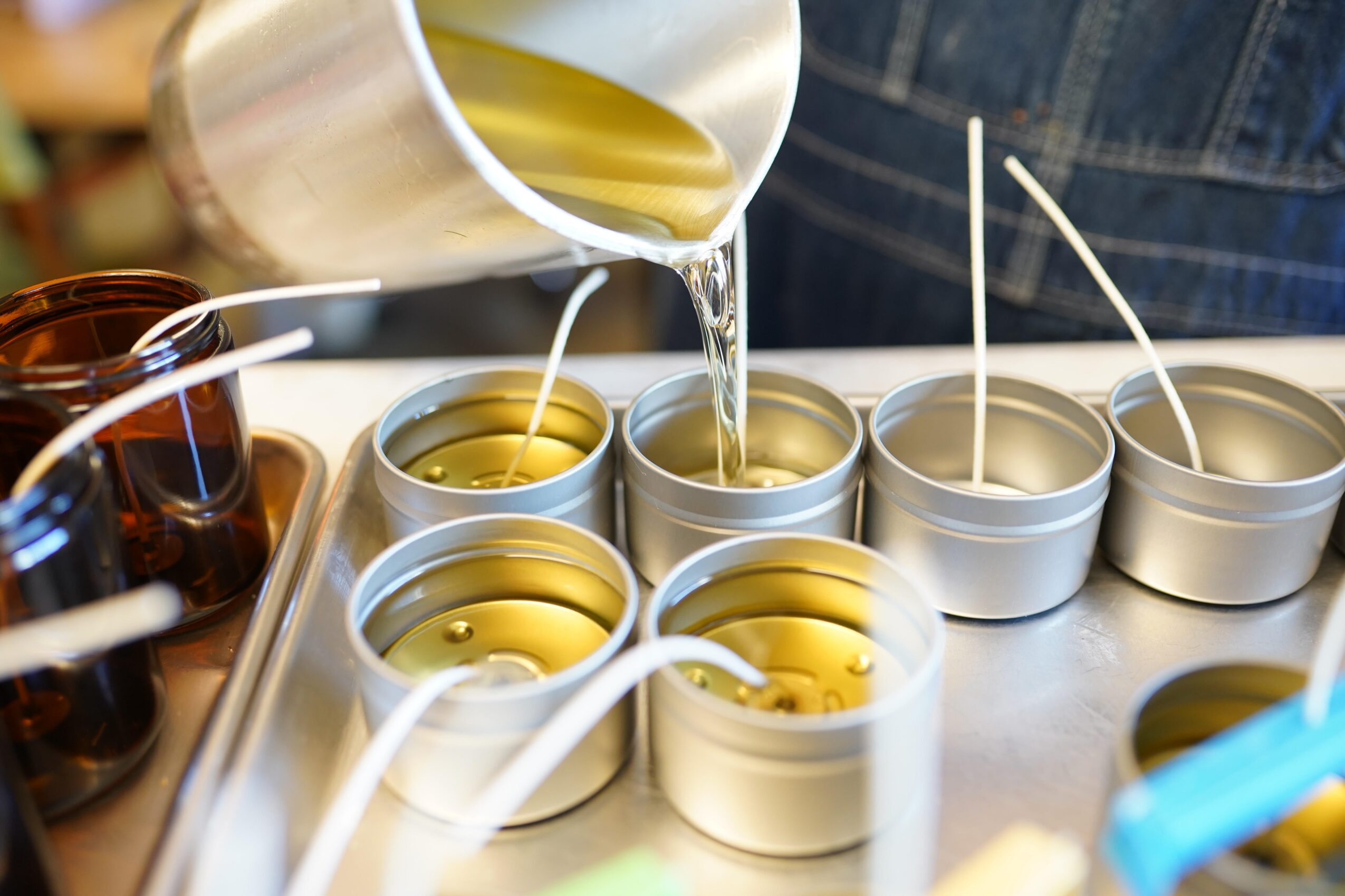 Person pouring yellow liquid into small white containers on a tray, with other containers and a denim-clad arm visible.